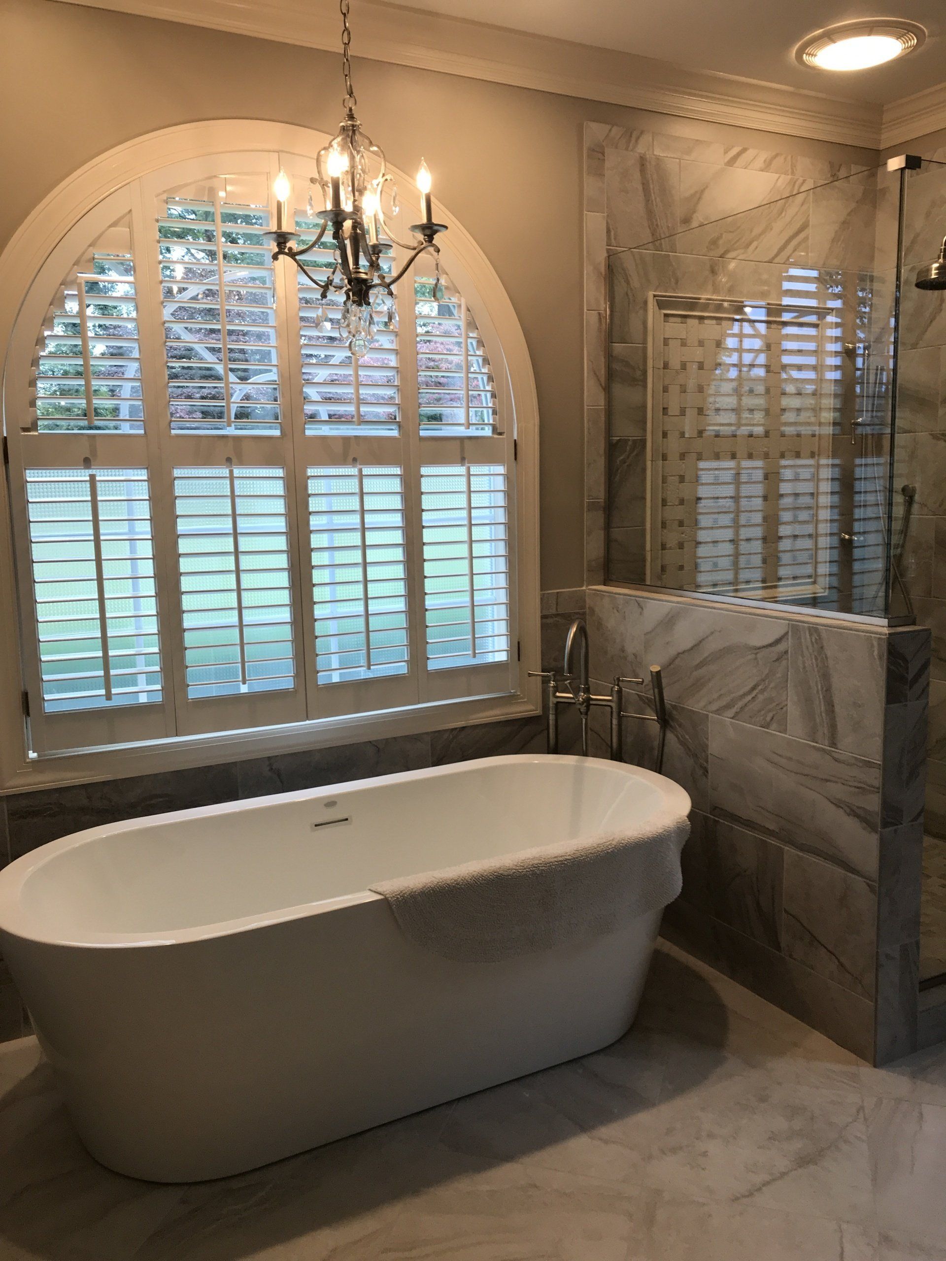 Elegant bathroom with a white freestanding tub, arched window with shutters, and marble walls.
