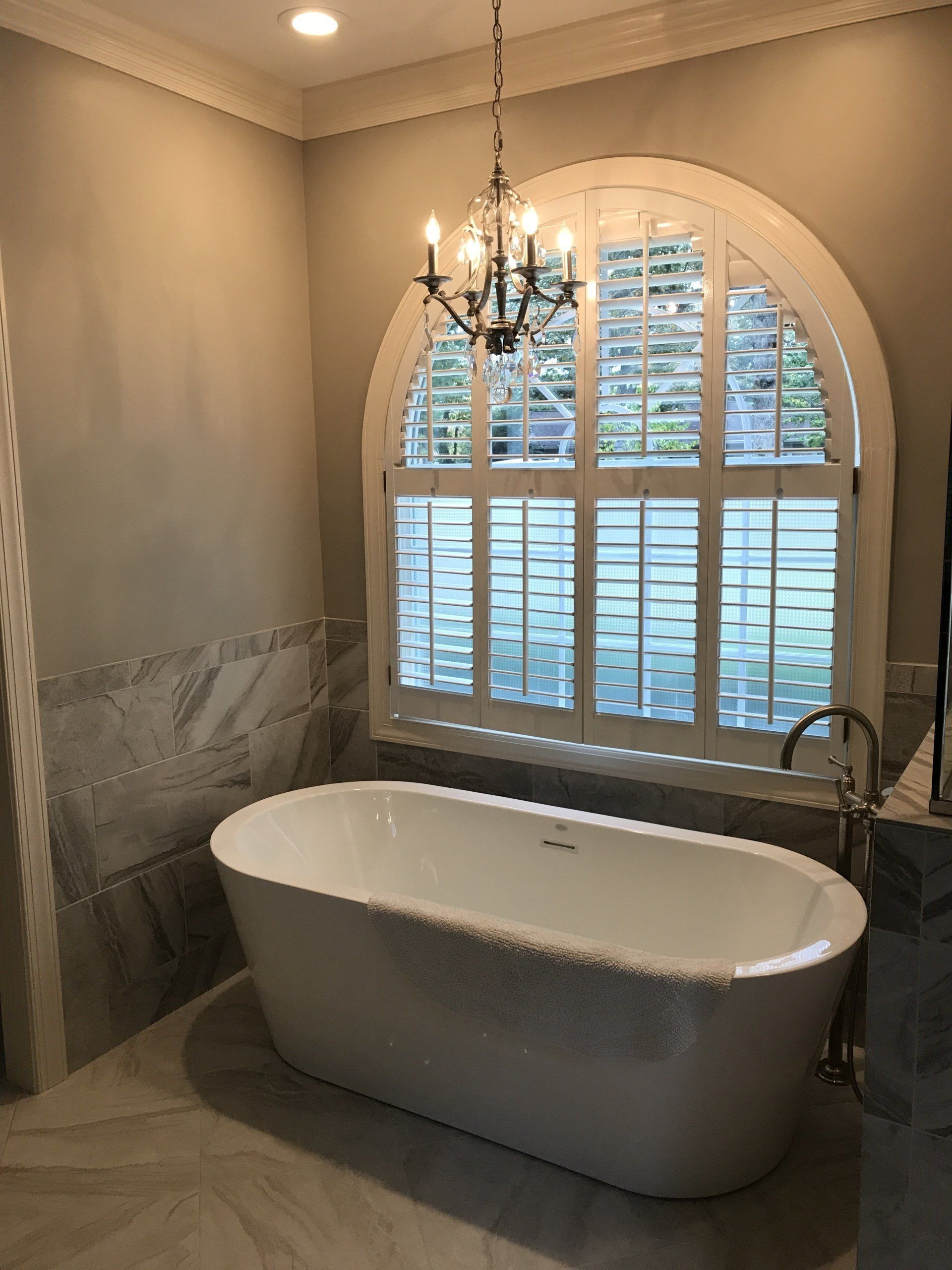 Bathroom with white oval tub, arched window with shutters, and chandelier.