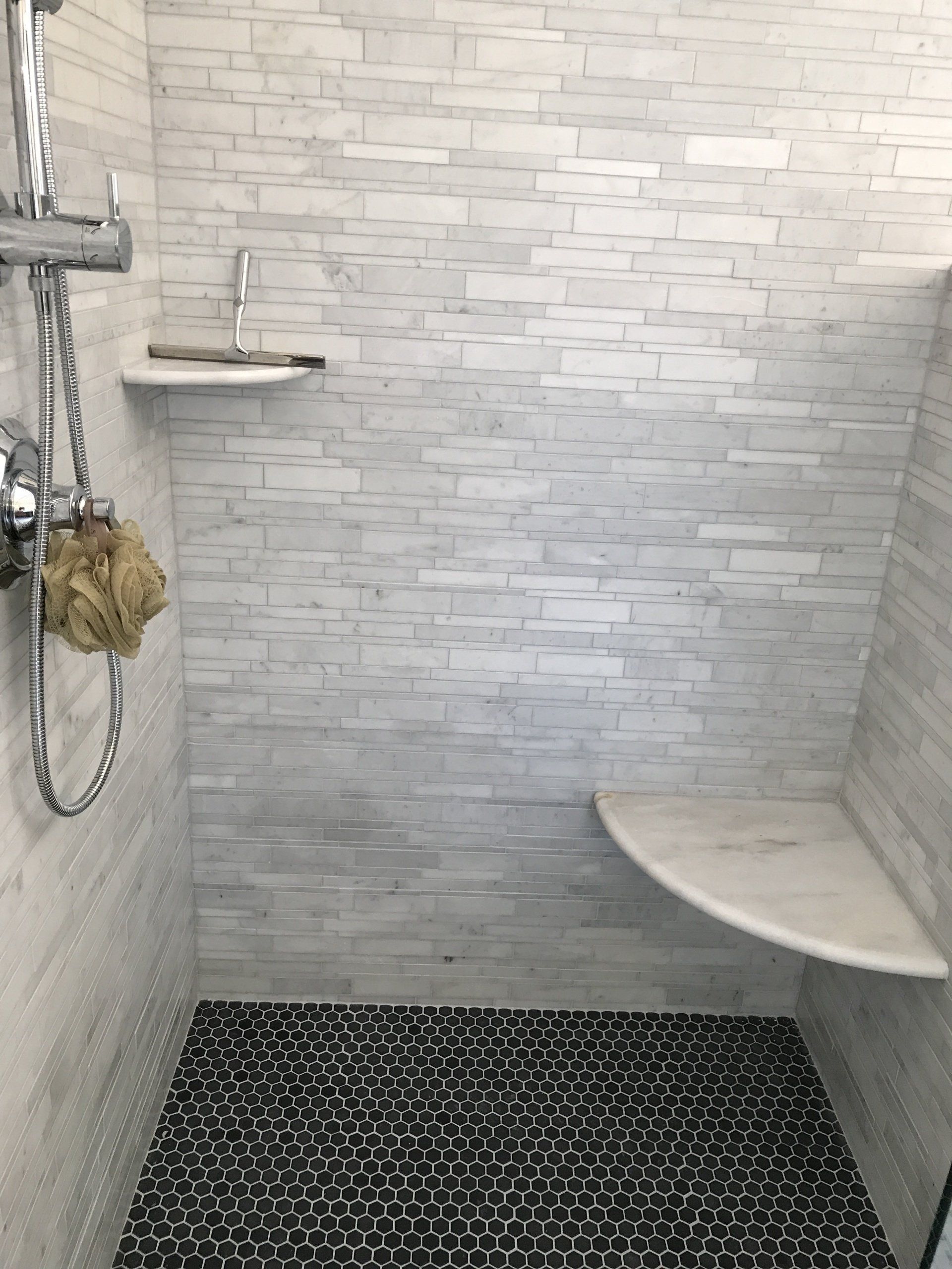Shower with white brick-like tile, gray and white rug, chrome fixtures, and two white corner shelves.