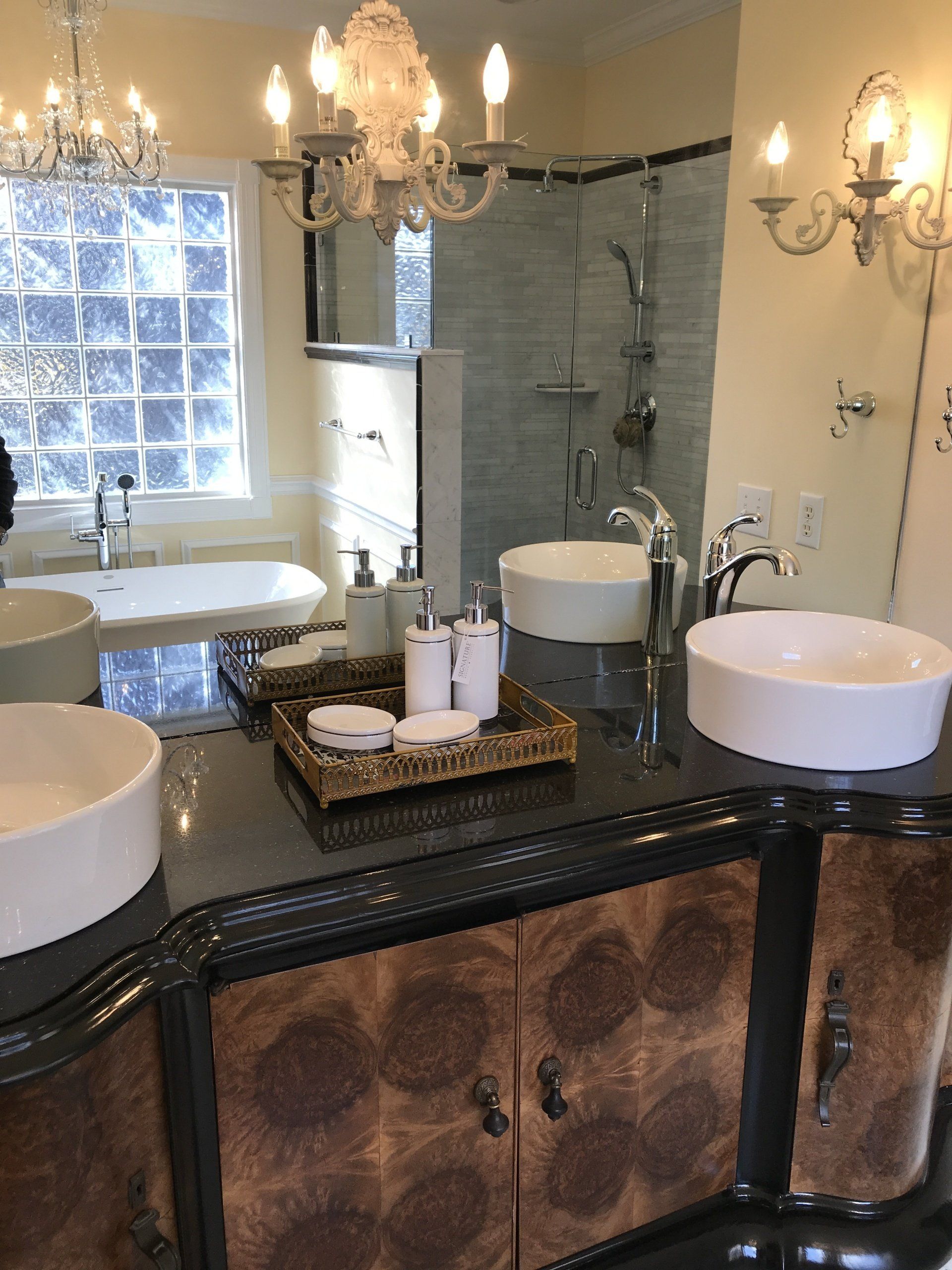 Bathroom with double sinks, vanity, and shower.  Cream walls, dark countertop, ornate cabinetry, chandelier, and glass block window.