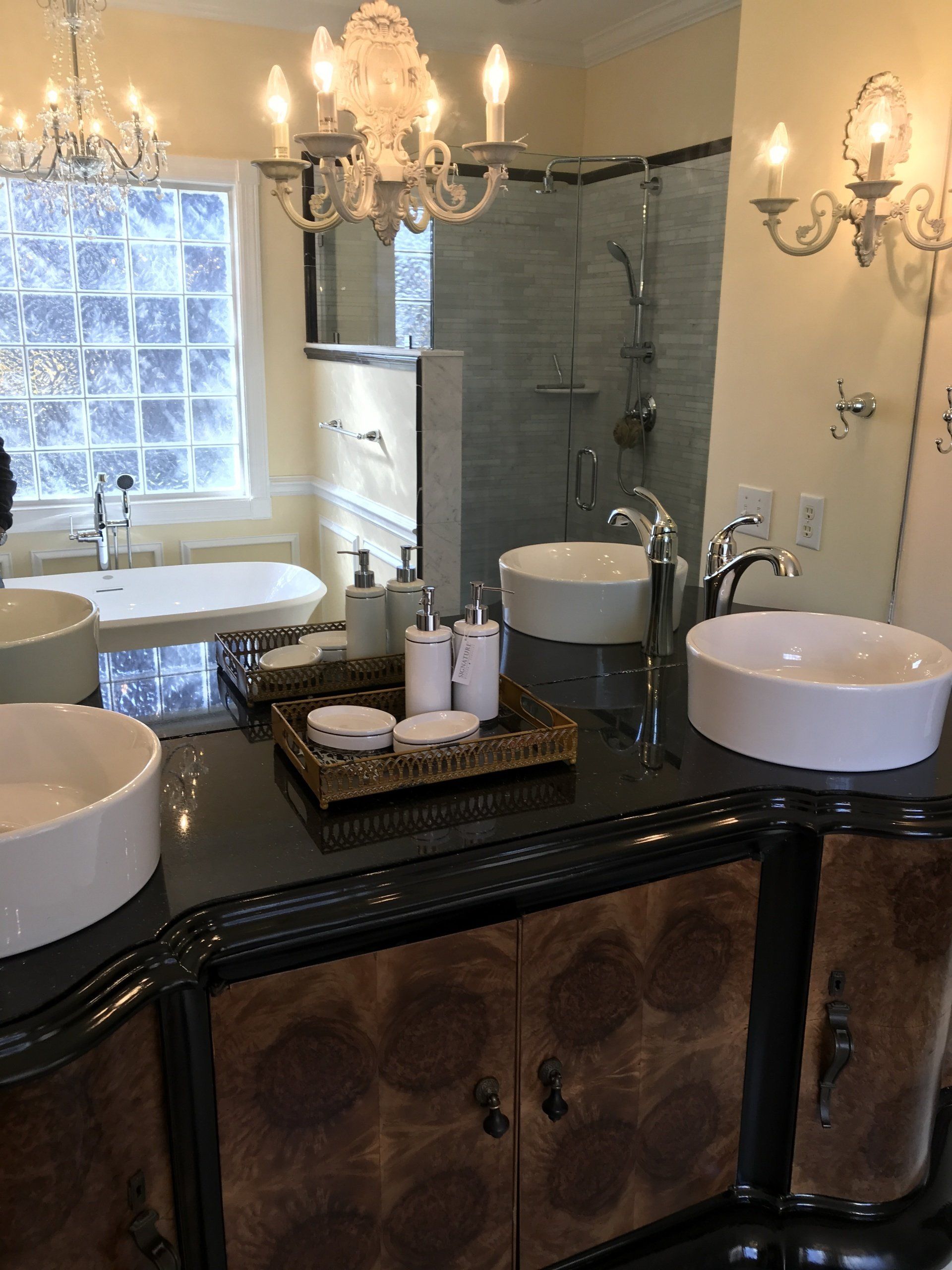 Bathroom vanity with two white sinks, black countertop, ornate wood cabinet, glass shower, chandelier.