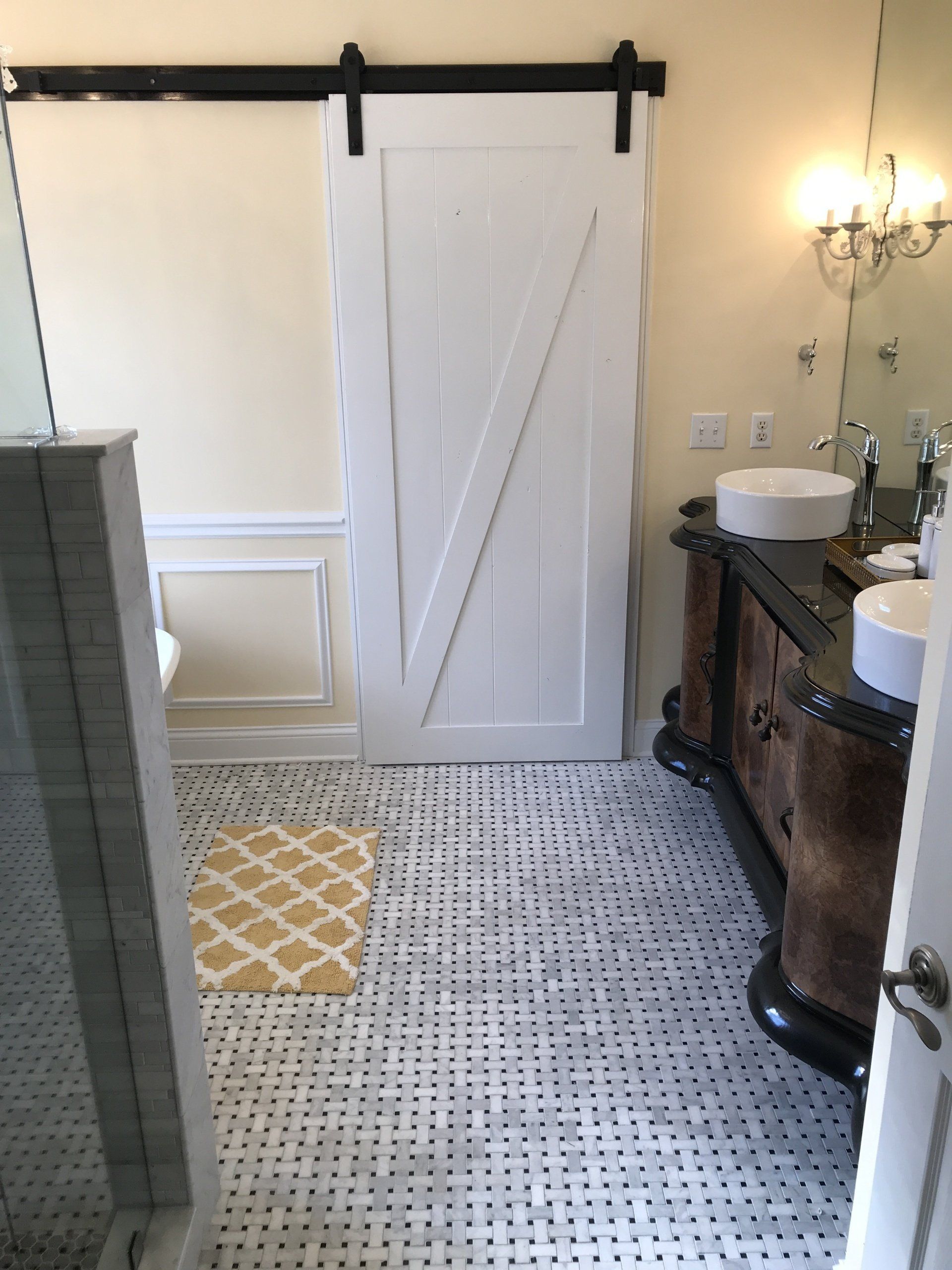 Bathroom with white sliding barn door, vanity, and white and black tiled floor; a yellow rug is near the door.
