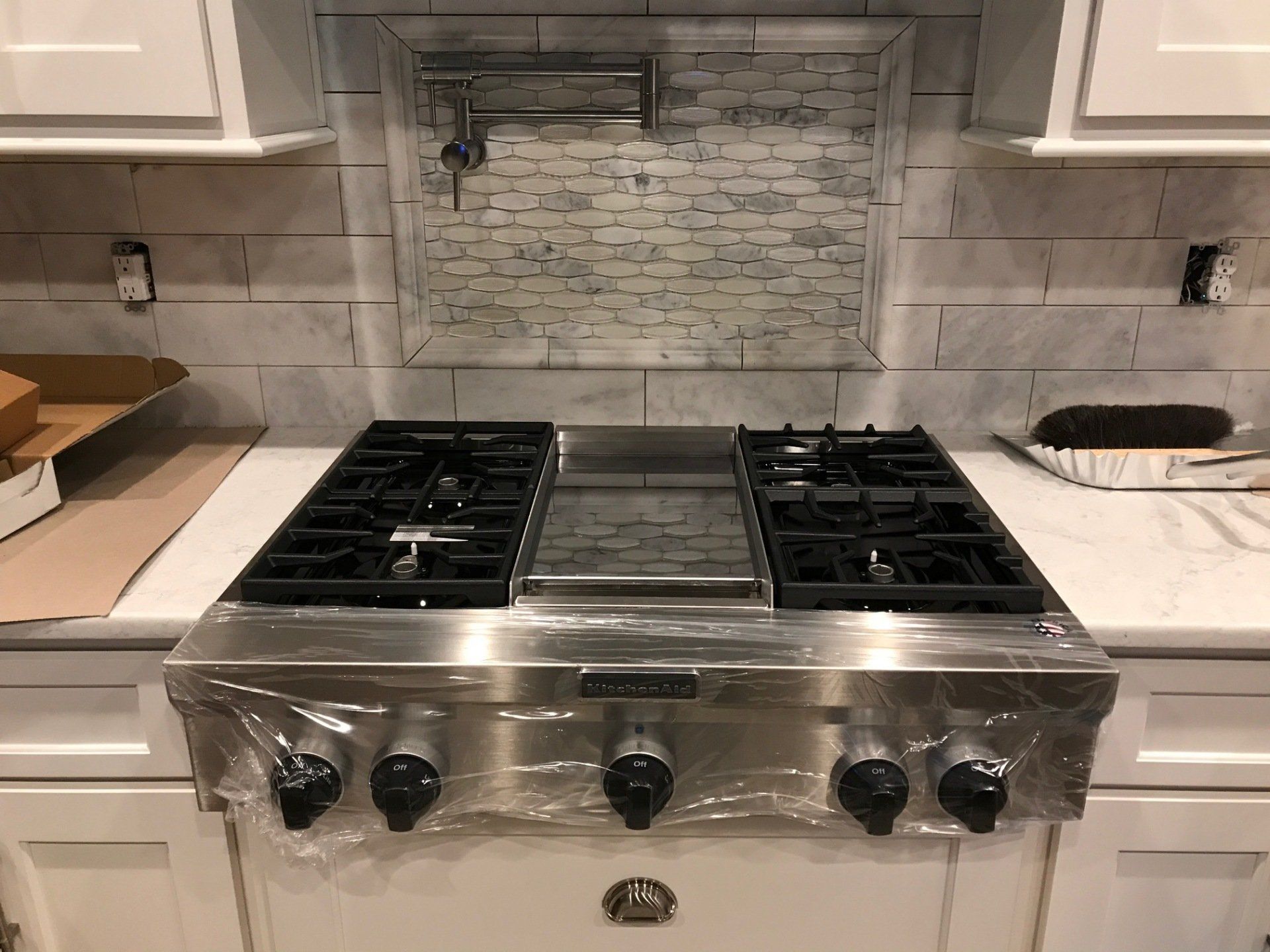 Stainless steel gas cooktop with knobs, backsplash, and white cabinets in a kitchen.