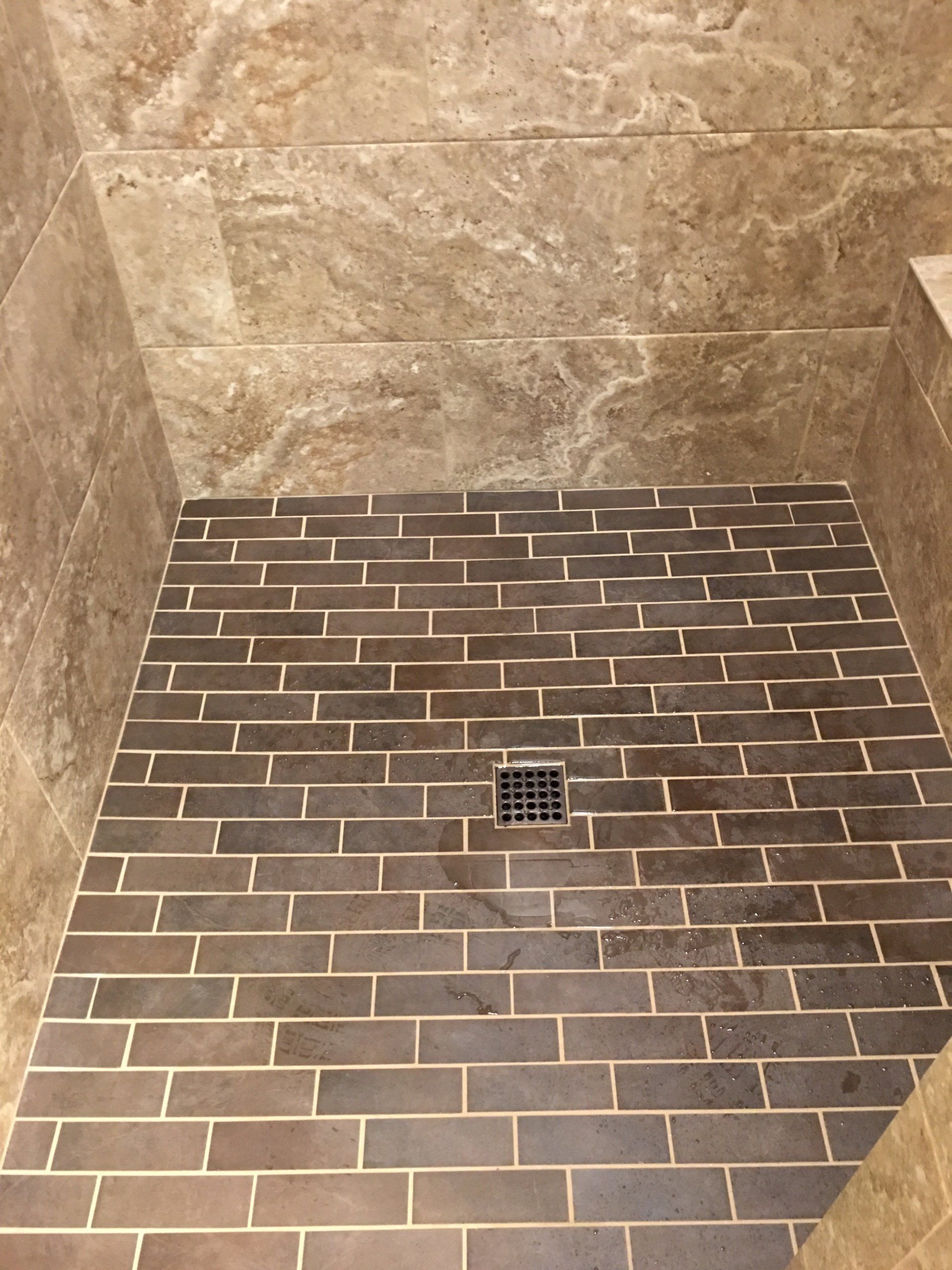 Shower stall with dark brick-like tiles on the floor and tan marble-look tiles on the walls. Drain visible in the center.