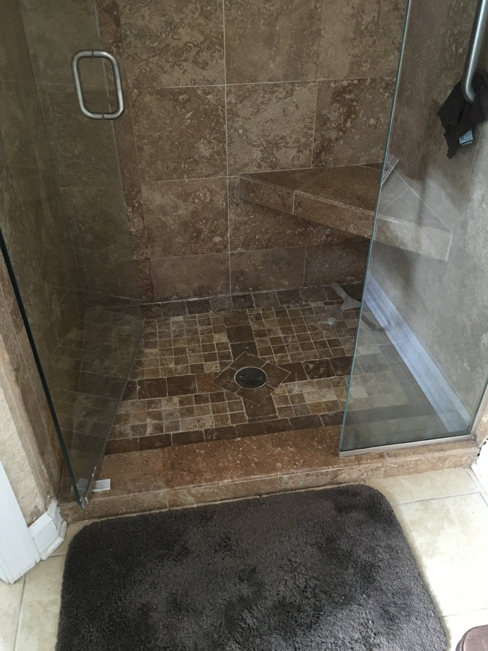 Shower with brown tile and glass door; a dark bath mat sits outside.