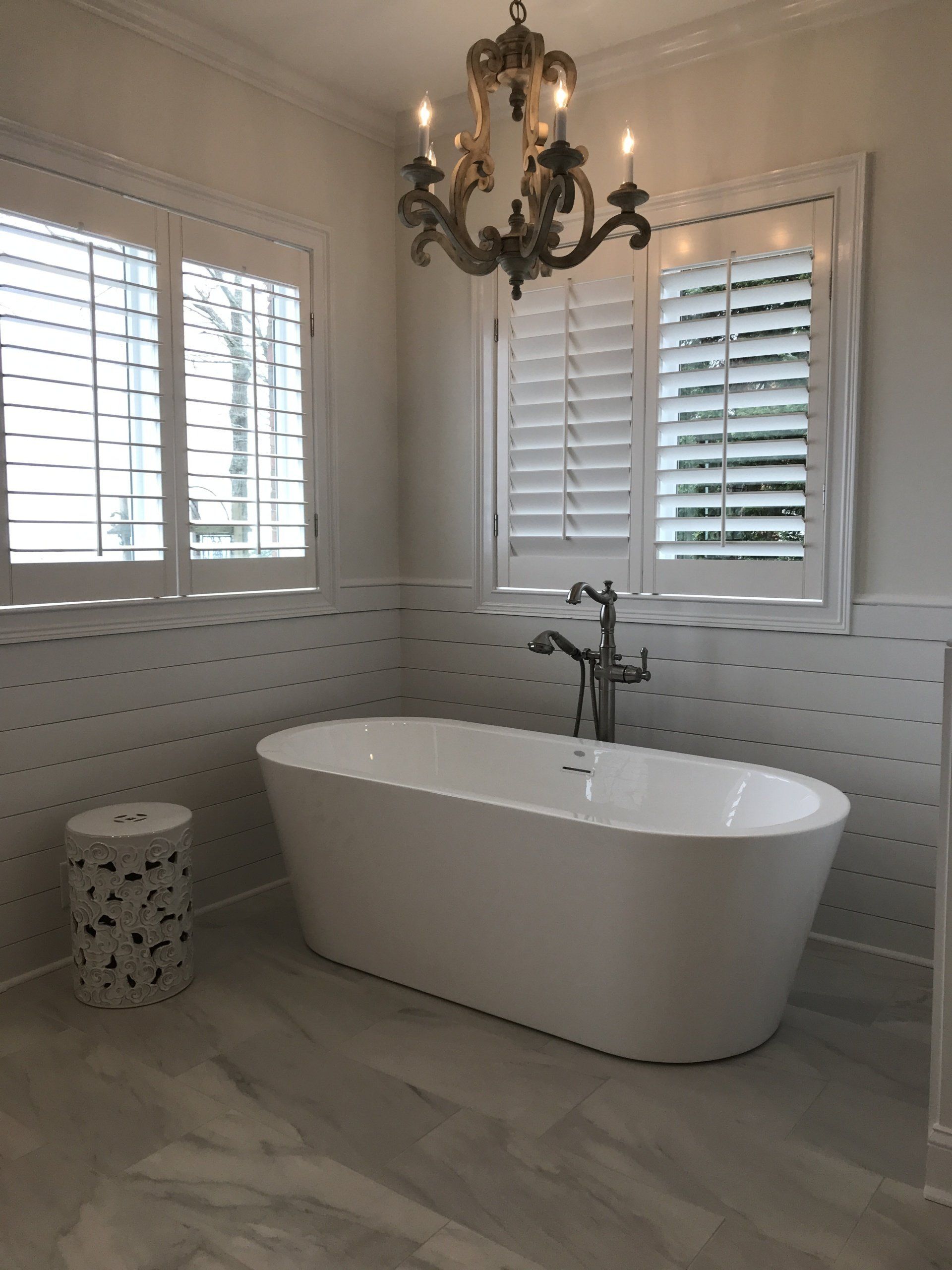 Bathroom with a white oval tub, shutters, and a decorative chandelier.