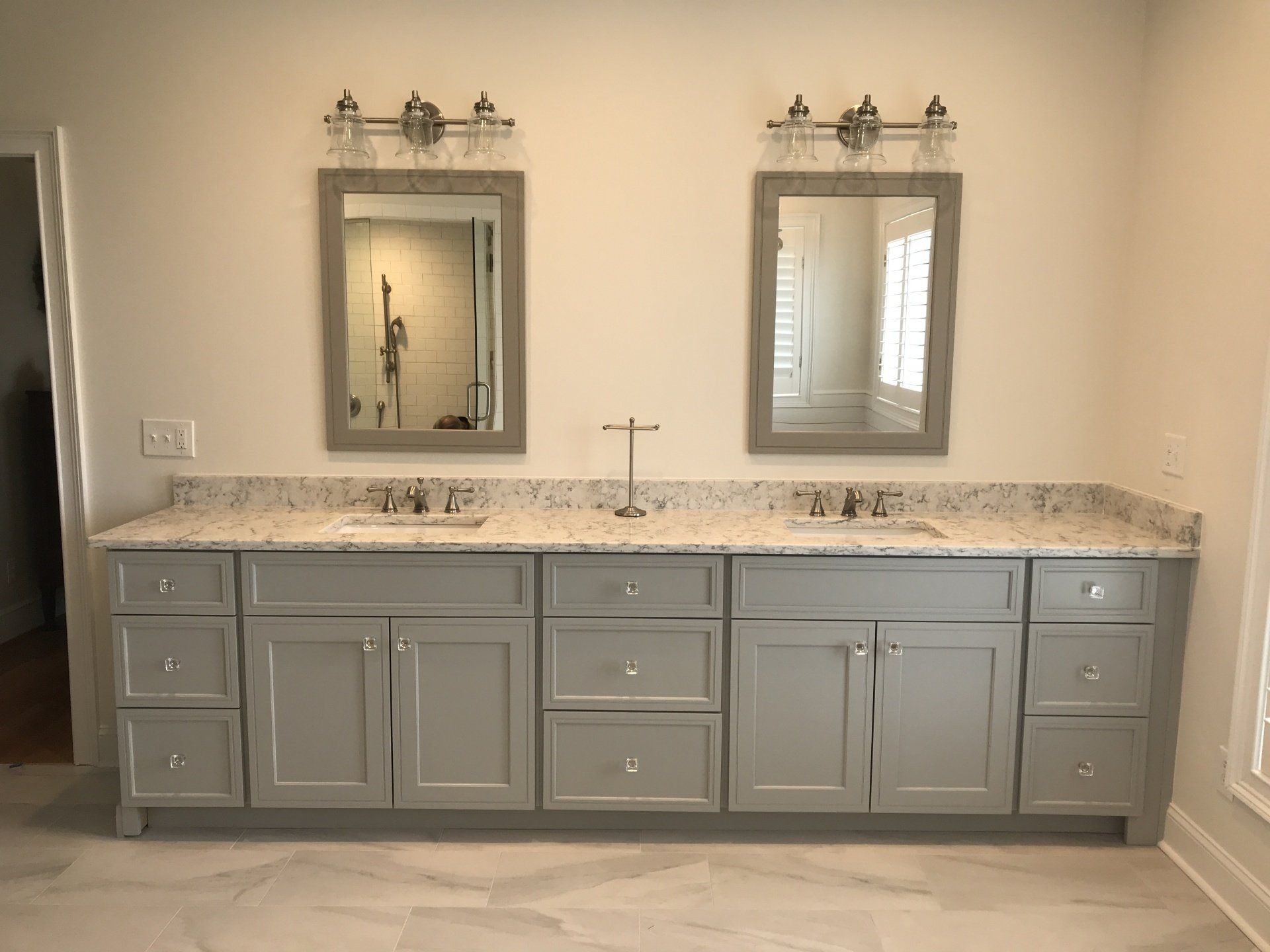 Bathroom with gray cabinets, two mirrors, and a light-colored countertop. Two sinks and a jewelry holder are visible.