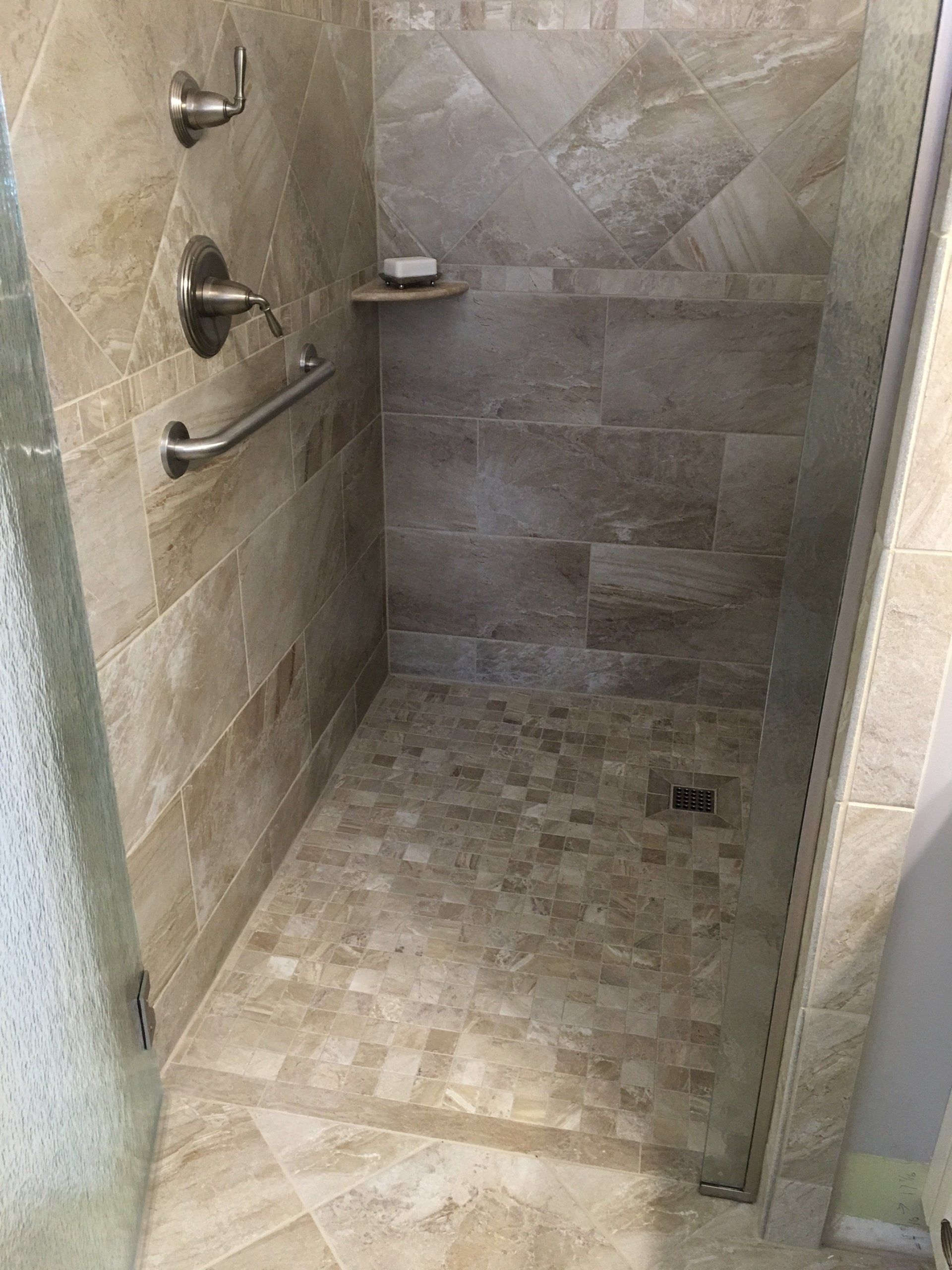 Walk-in shower with beige tile walls and floor, grab bar, built-in soap dish, and glass door.