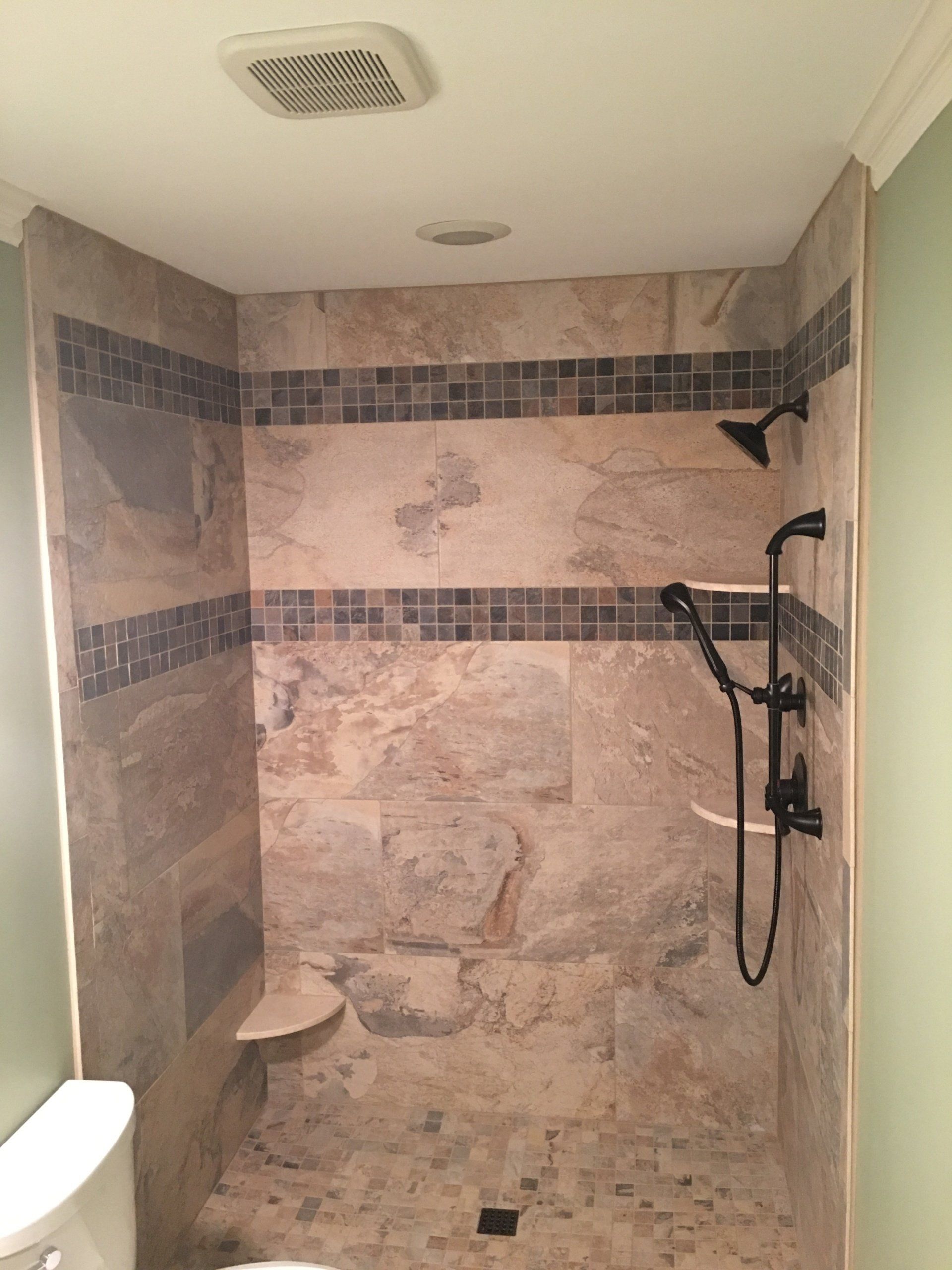 Shower with beige tiles, dark accents, shower heads, and a corner seat; above a toilet.
