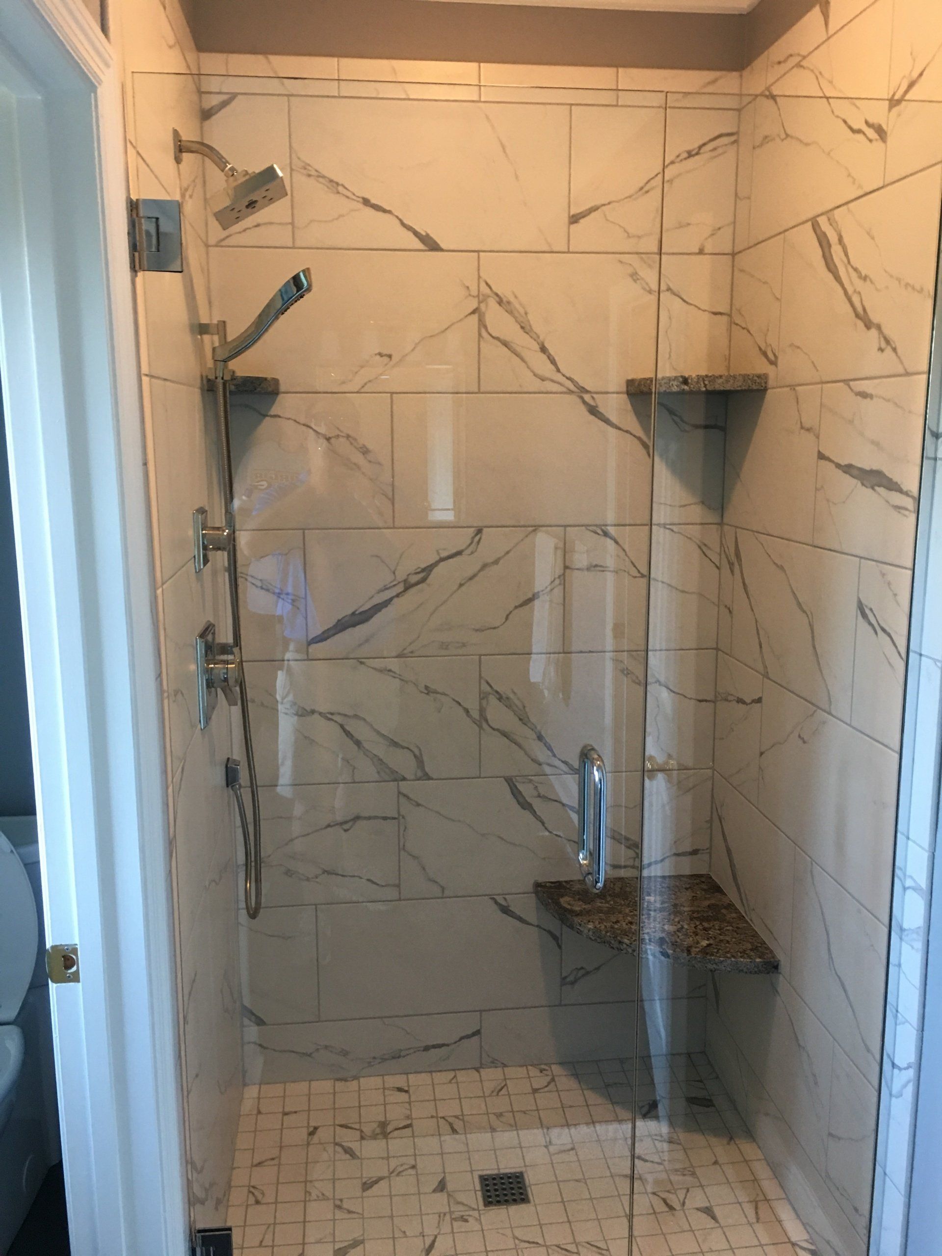Modern shower with marble-like tiled walls, glass door, and built-in seat; chrome fixtures.