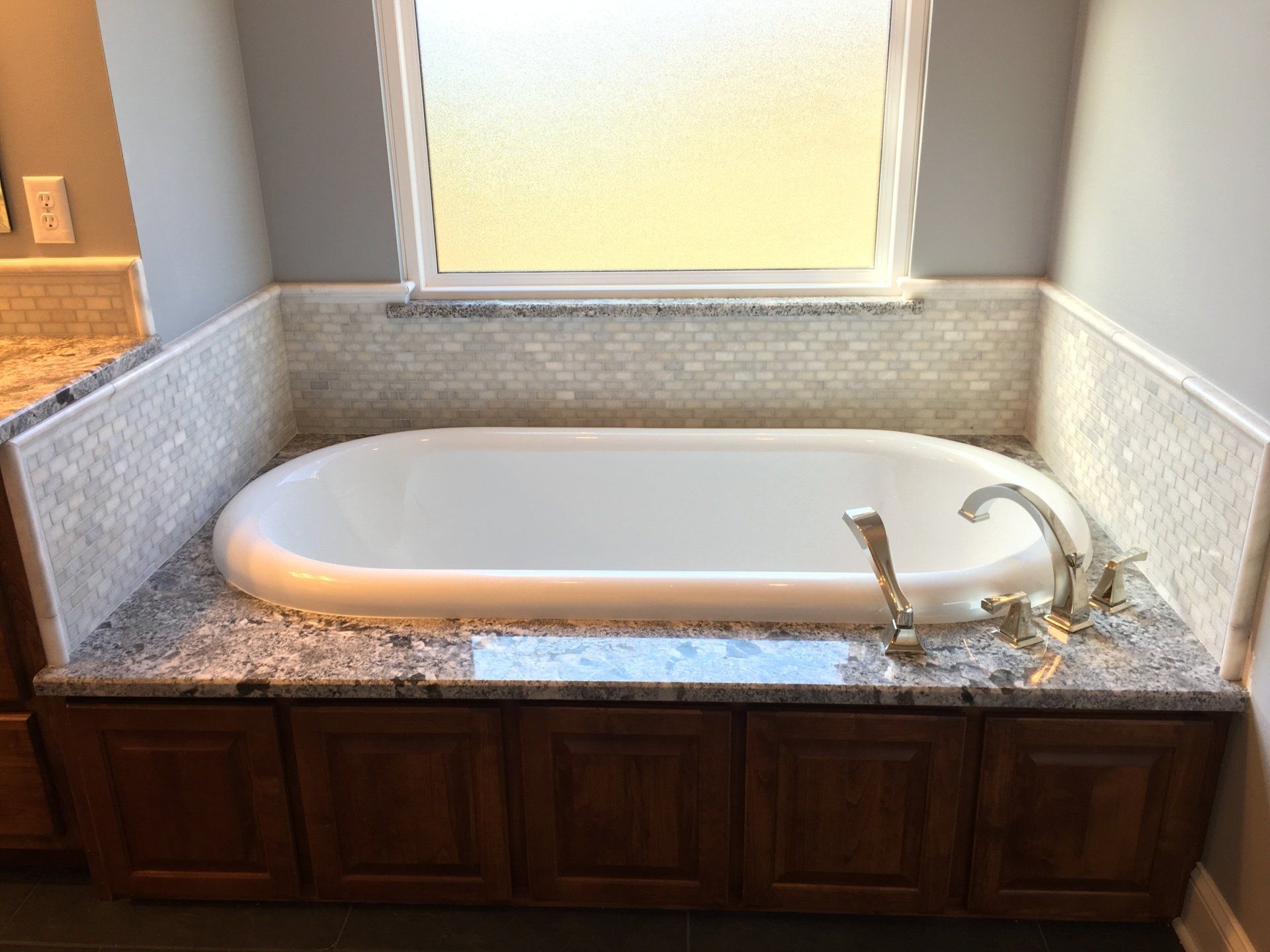 Bathroom with oval bathtub set in a granite countertop. Cabinetry below, mosaic tile backsplash, and a window above.