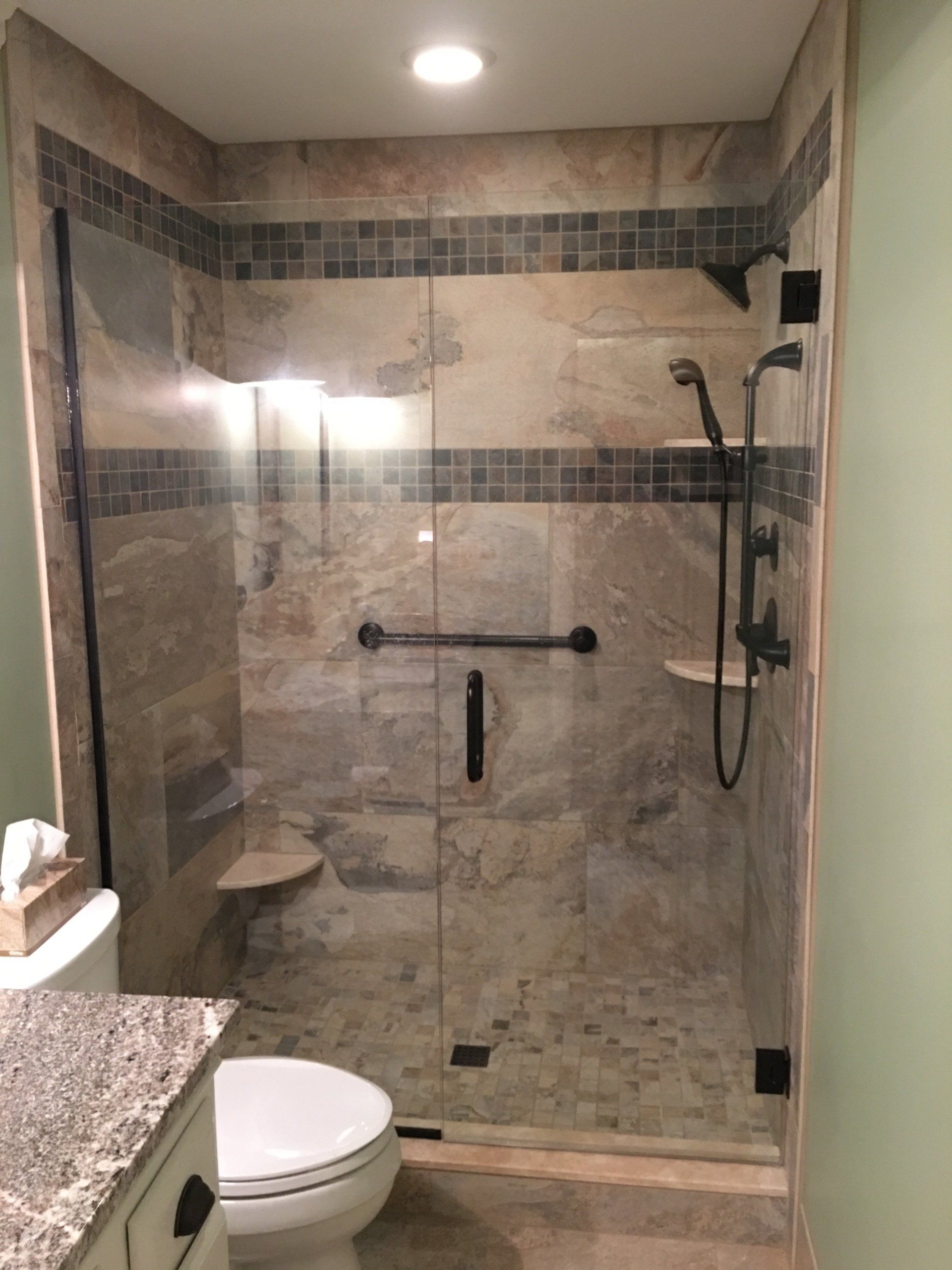 A tiled walk-in shower with glass doors, grab bar, and built-in bench, with a toilet in the foreground.