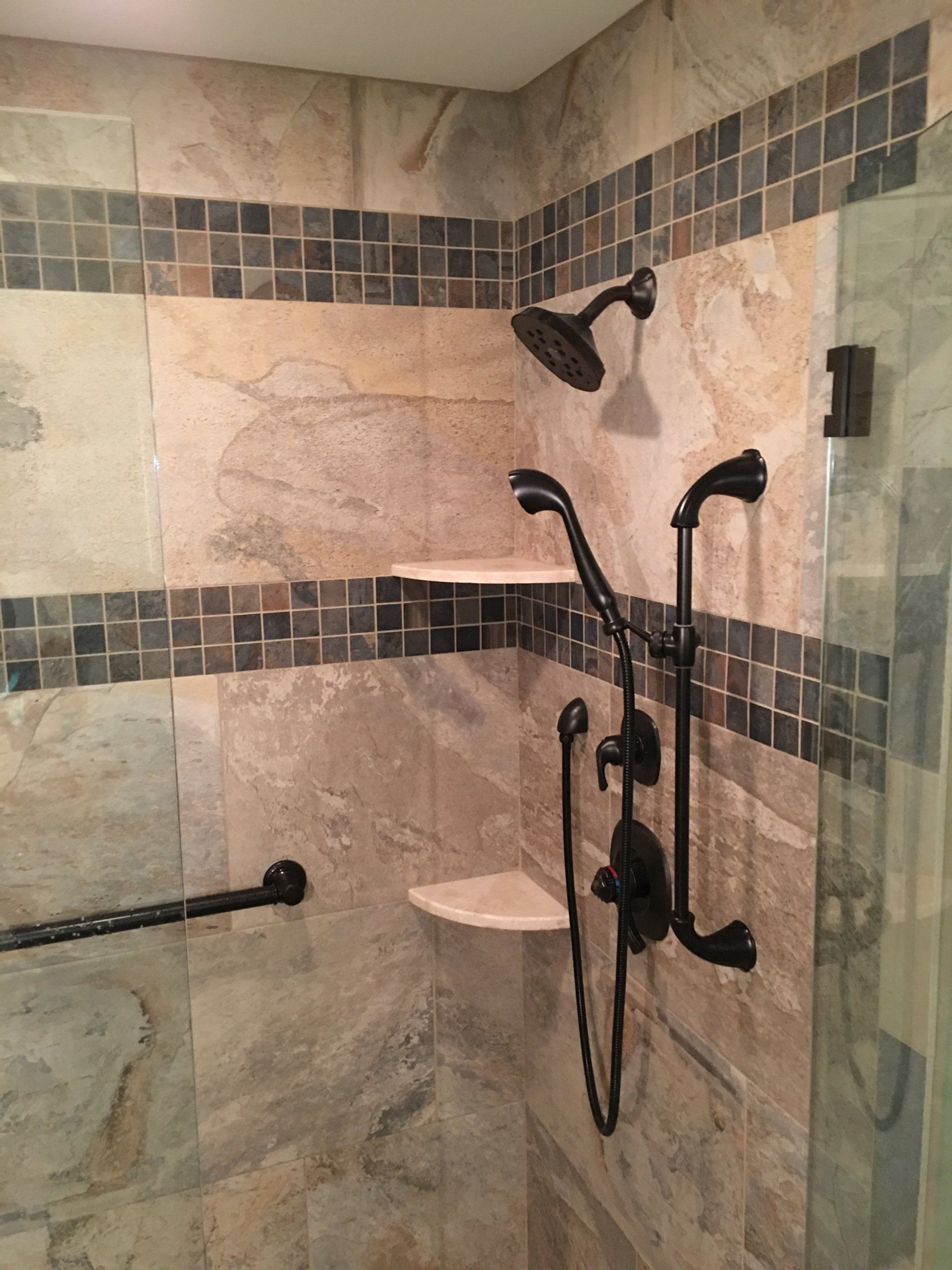 Shower with brown and beige stone tile, dark fixtures, shelves, and glass door.