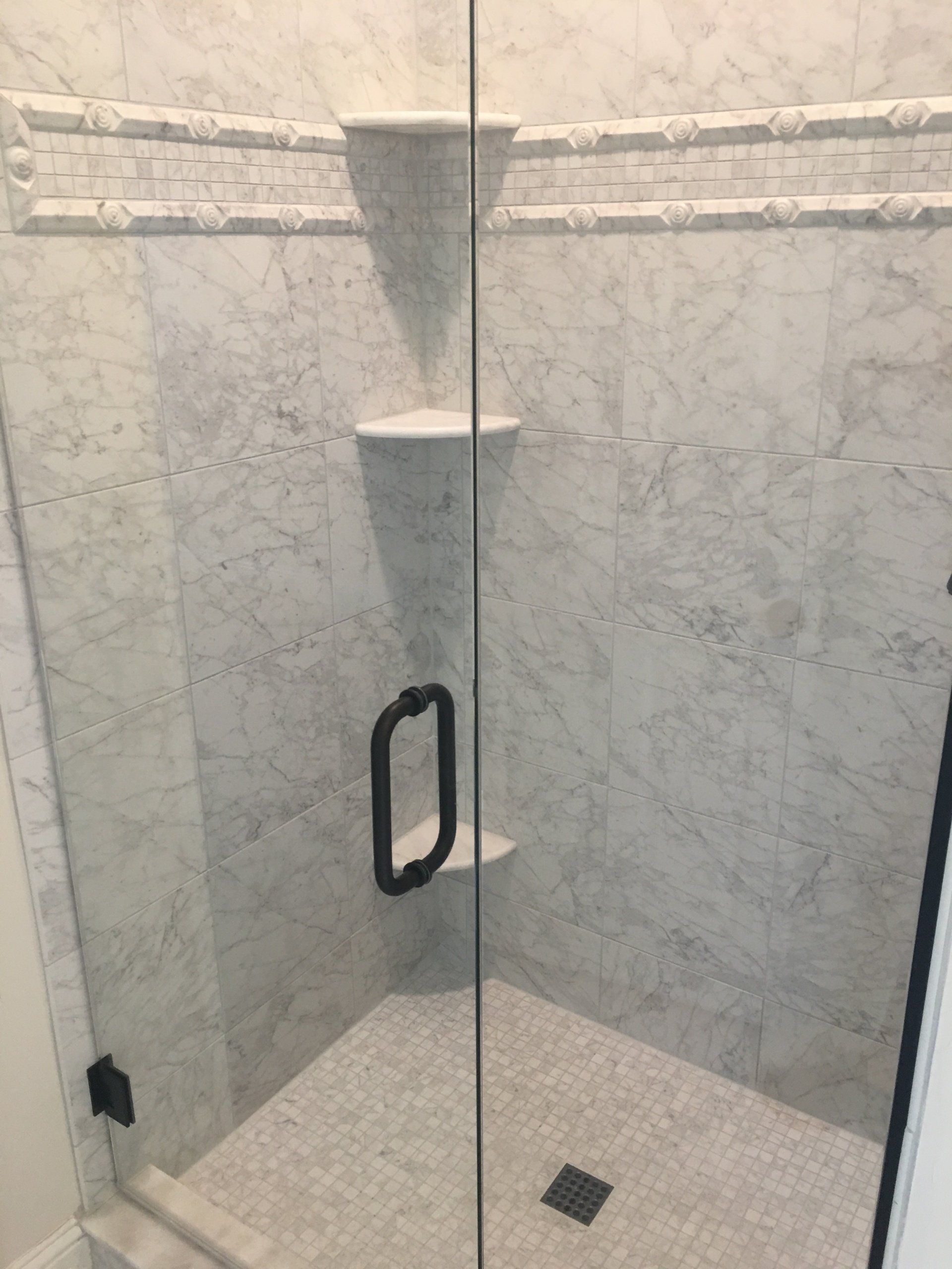 A marble-tiled shower with a glass door, black handle, and two built-in shelves.