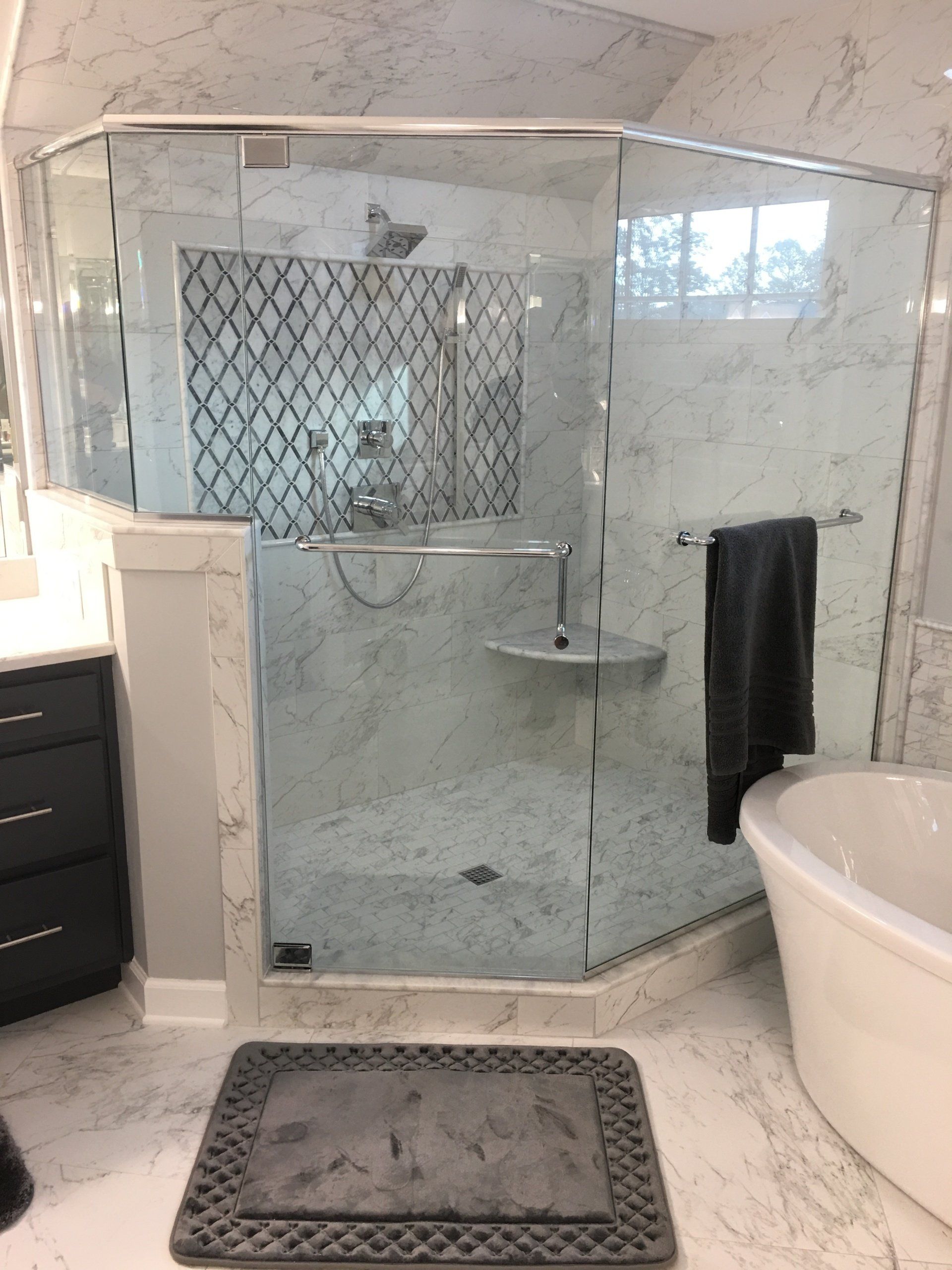A modern bathroom with a glass shower, marble walls, a dark gray towel, and a gray bathmat.