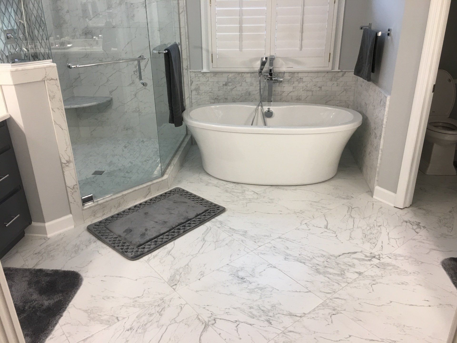 Modern bathroom with marble-look flooring, glass shower, white tub, and gray accents.