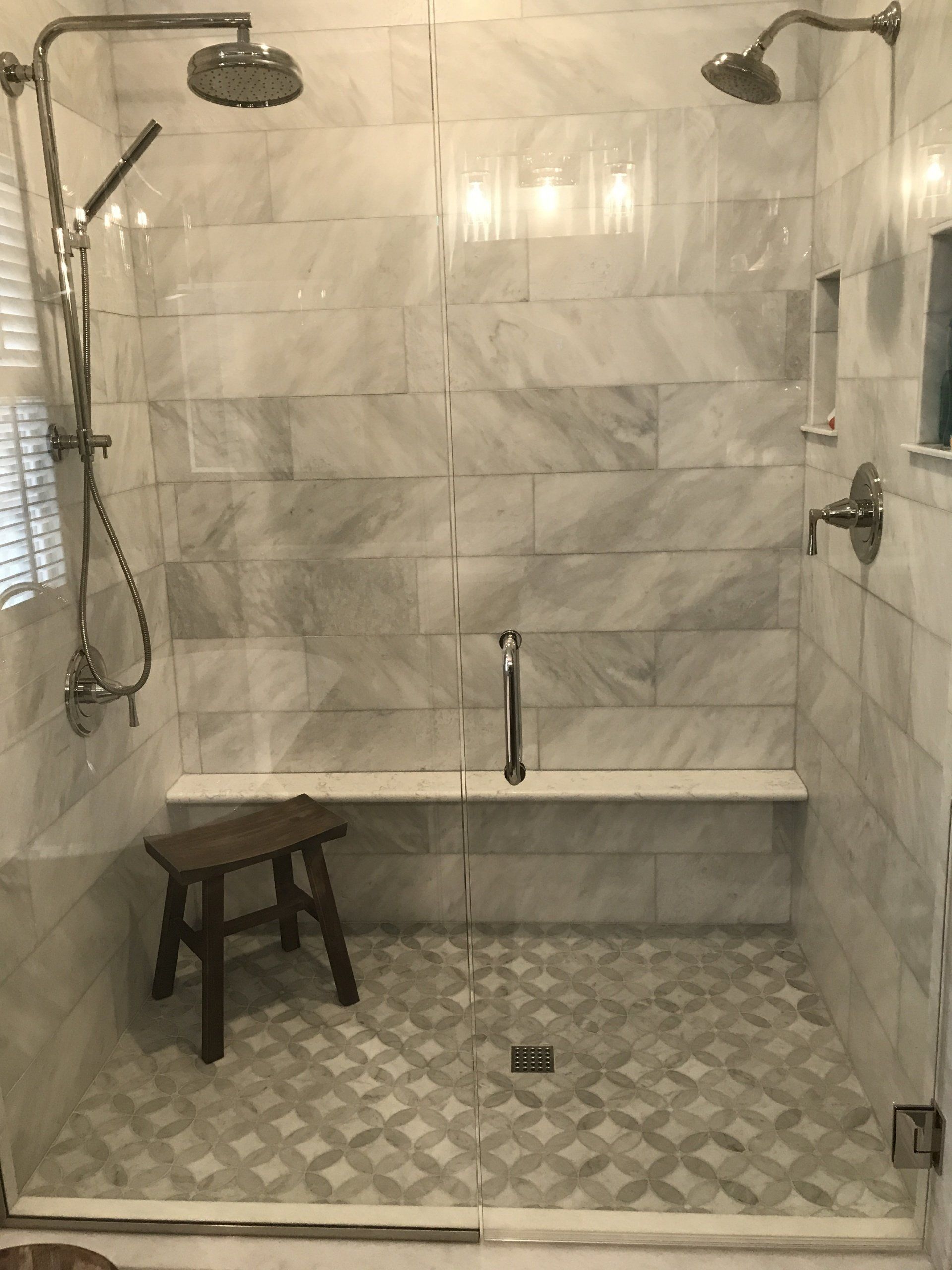 A marble-tiled shower with a seat and a stool. Two showerheads and a glass door.