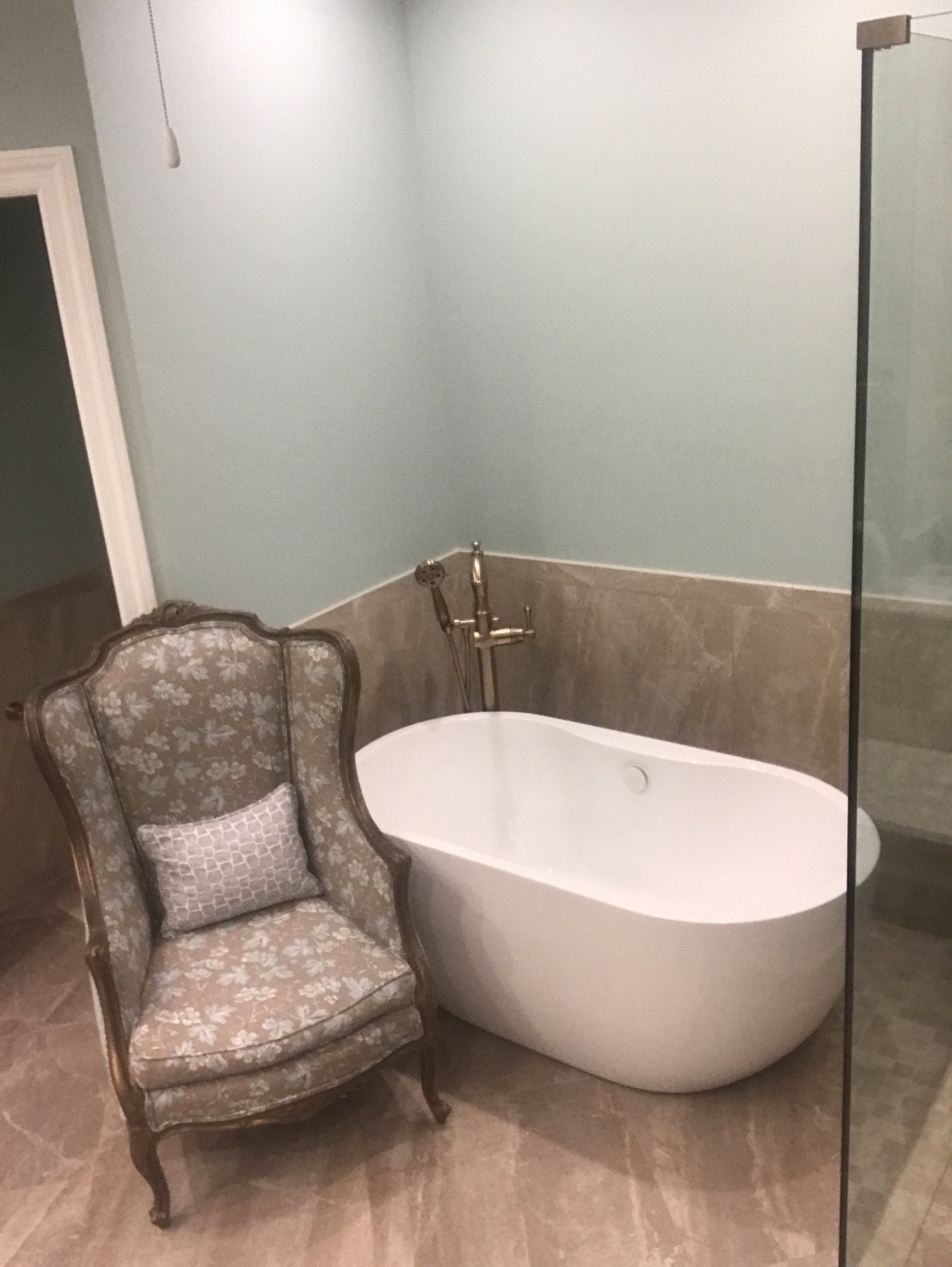 A luxurious bathroom with a white freestanding tub, antique chair, and glass shower.