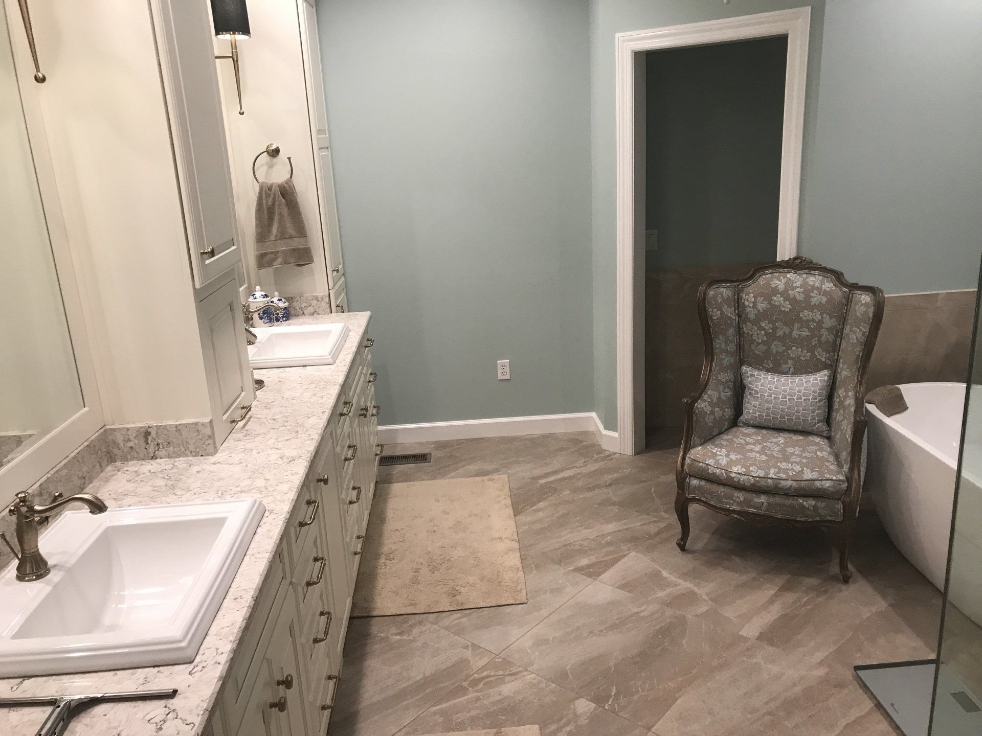Bathroom with double sinks, blue wall, patterned tile, and ornate armchair.