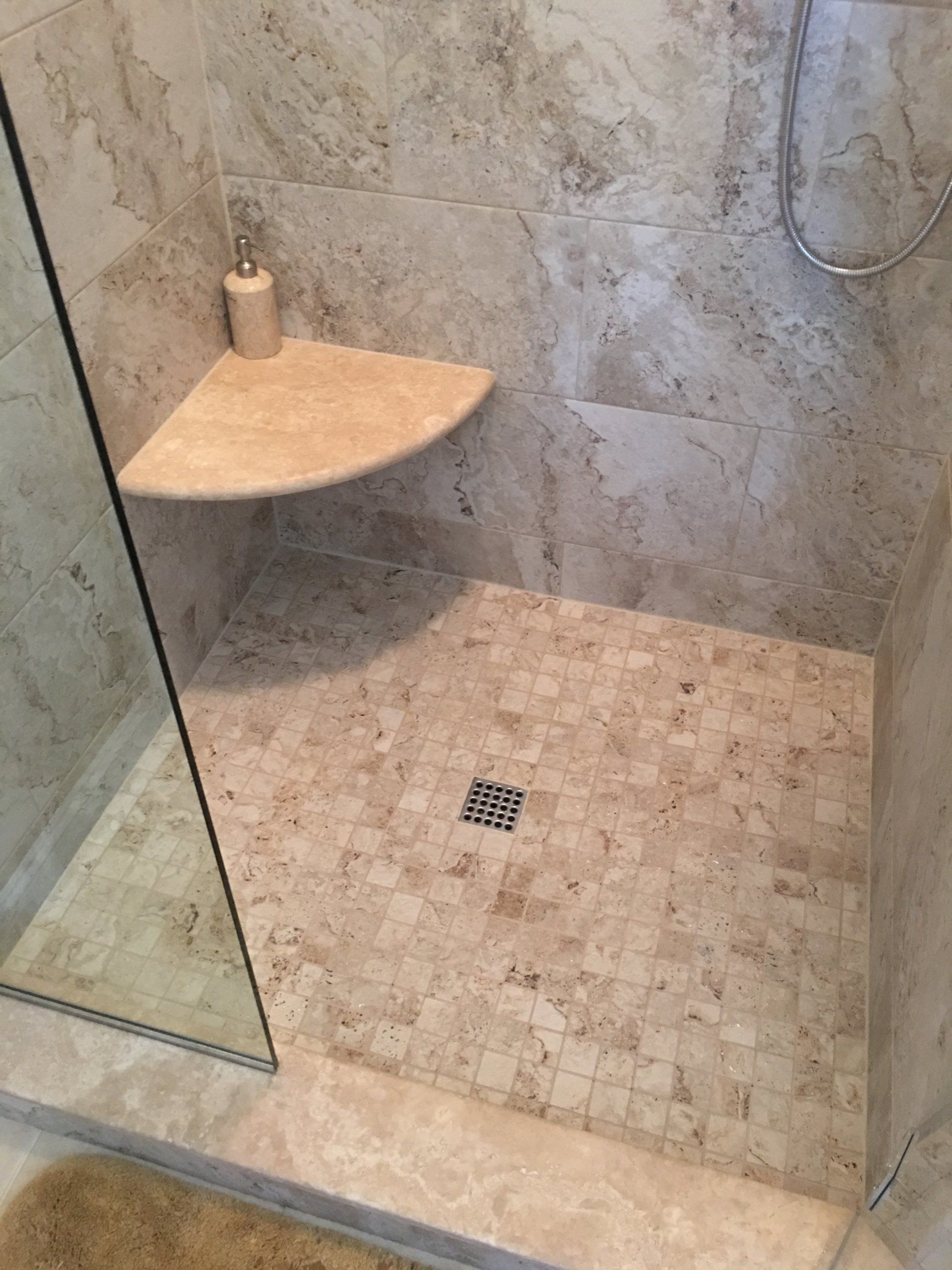 Shower with beige tile, corner shelf, and glass door; shower floor with a drain.