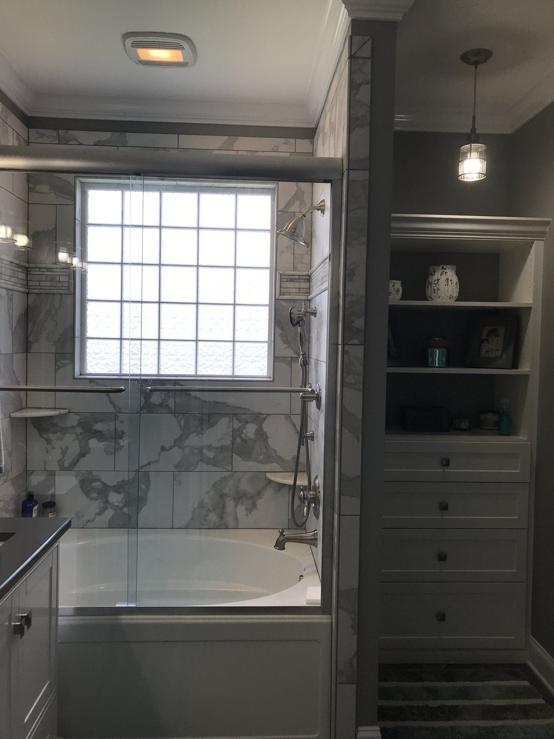 Bathroom with marble tile, a glass shower door, and built-in shelves; a window is centered above the tub.