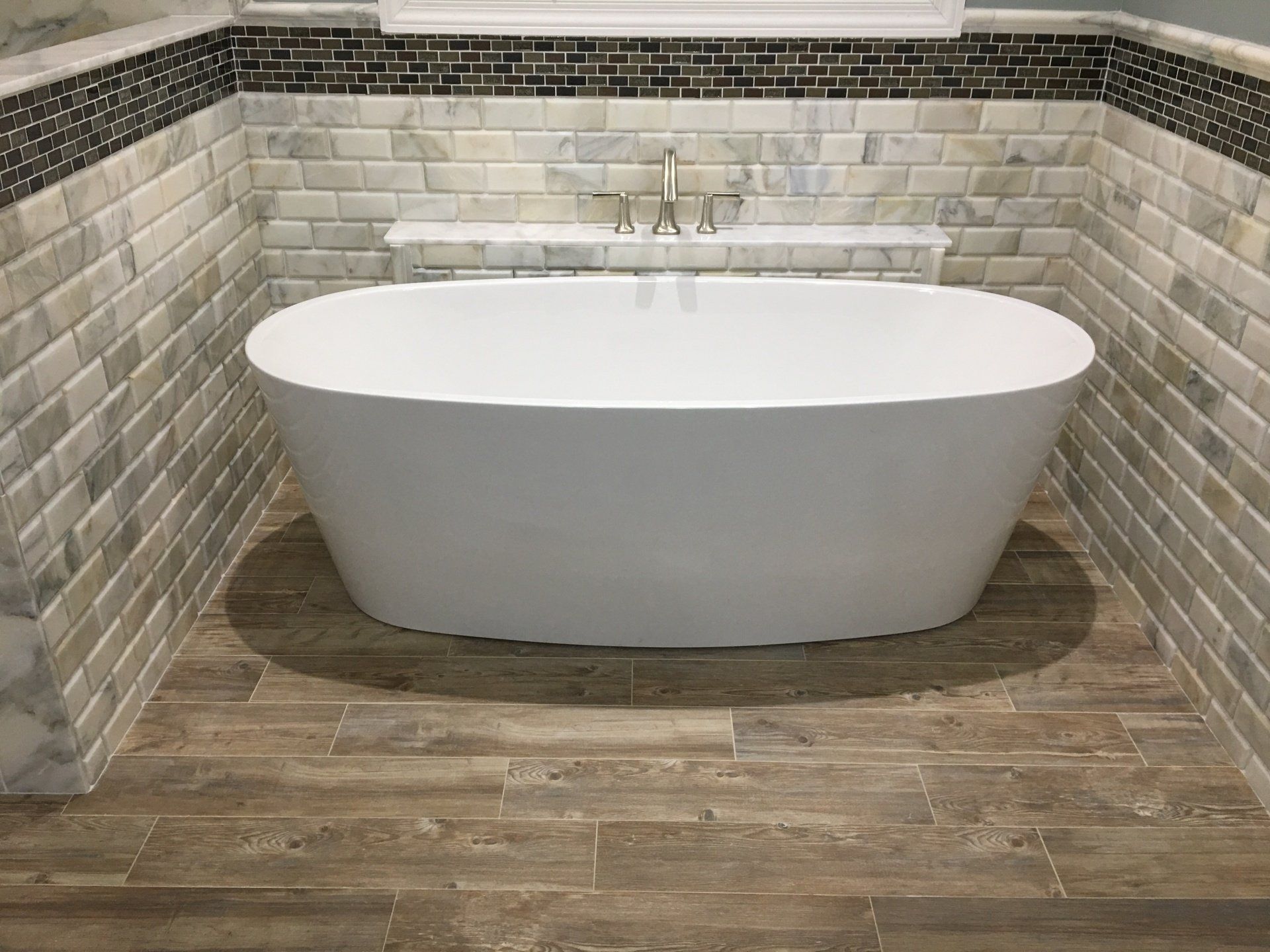 Freestanding white bathtub in a bathroom with wood-look floor tiles and marble-like wall tiles.