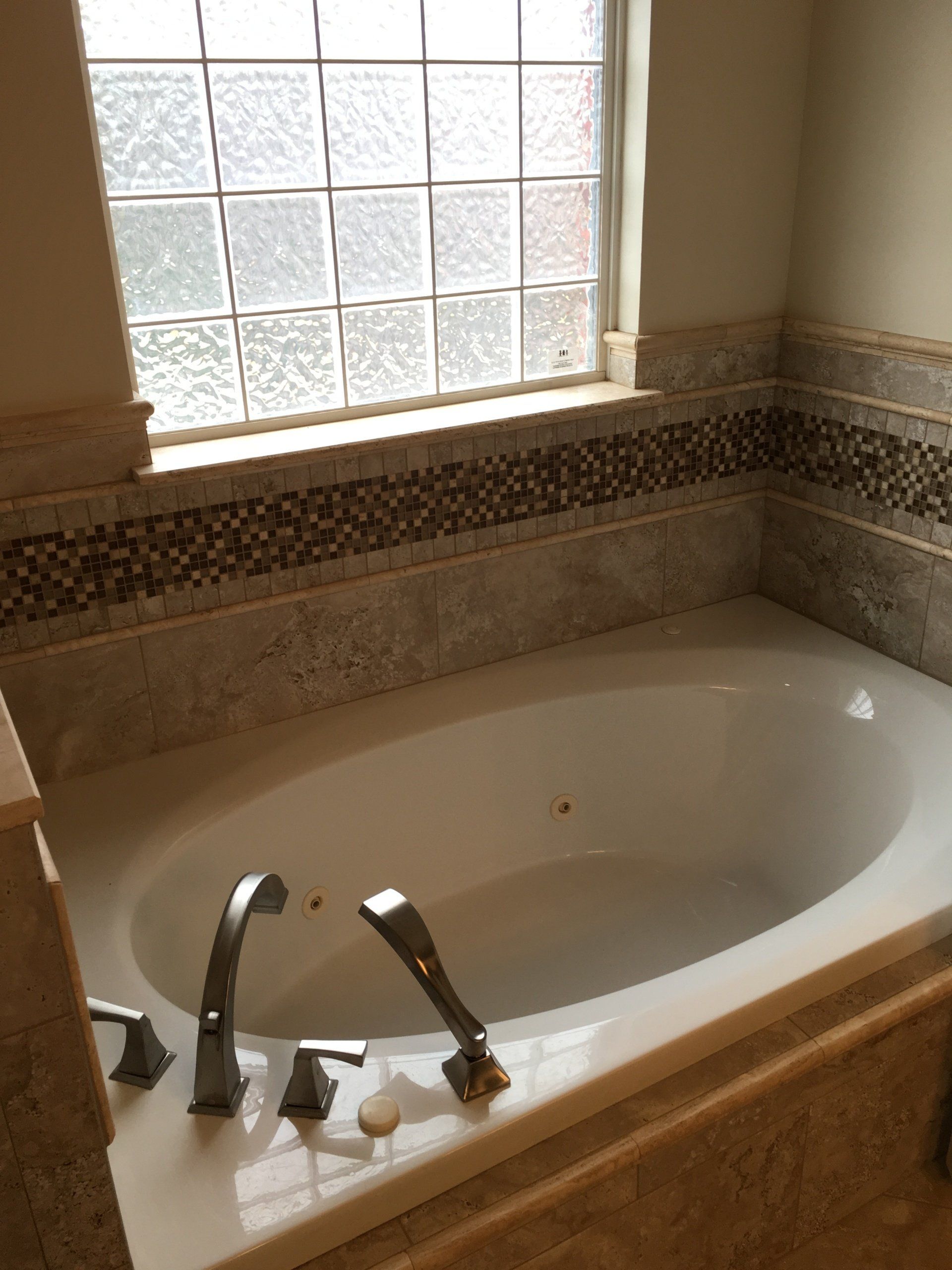 Oval bathtub with chrome fixtures, mosaic tile, and frosted window.