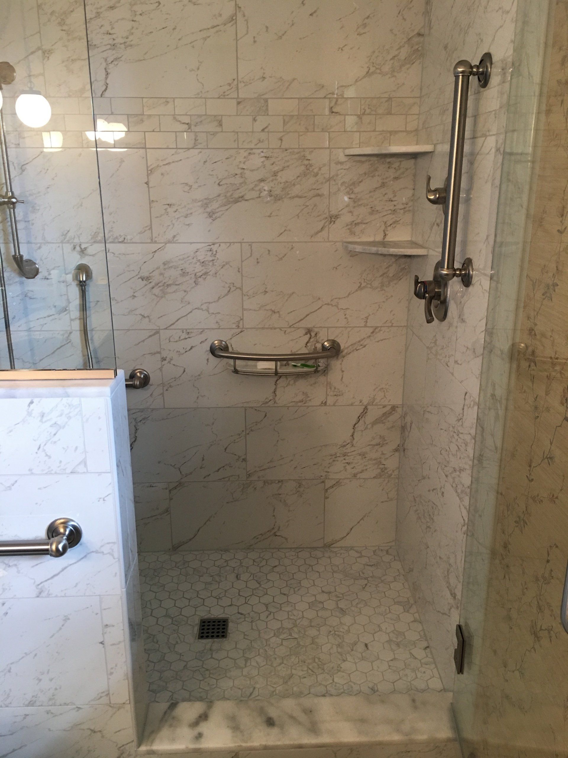 Marble-tiled accessible shower with grab bars and shelves. Clear glass door.