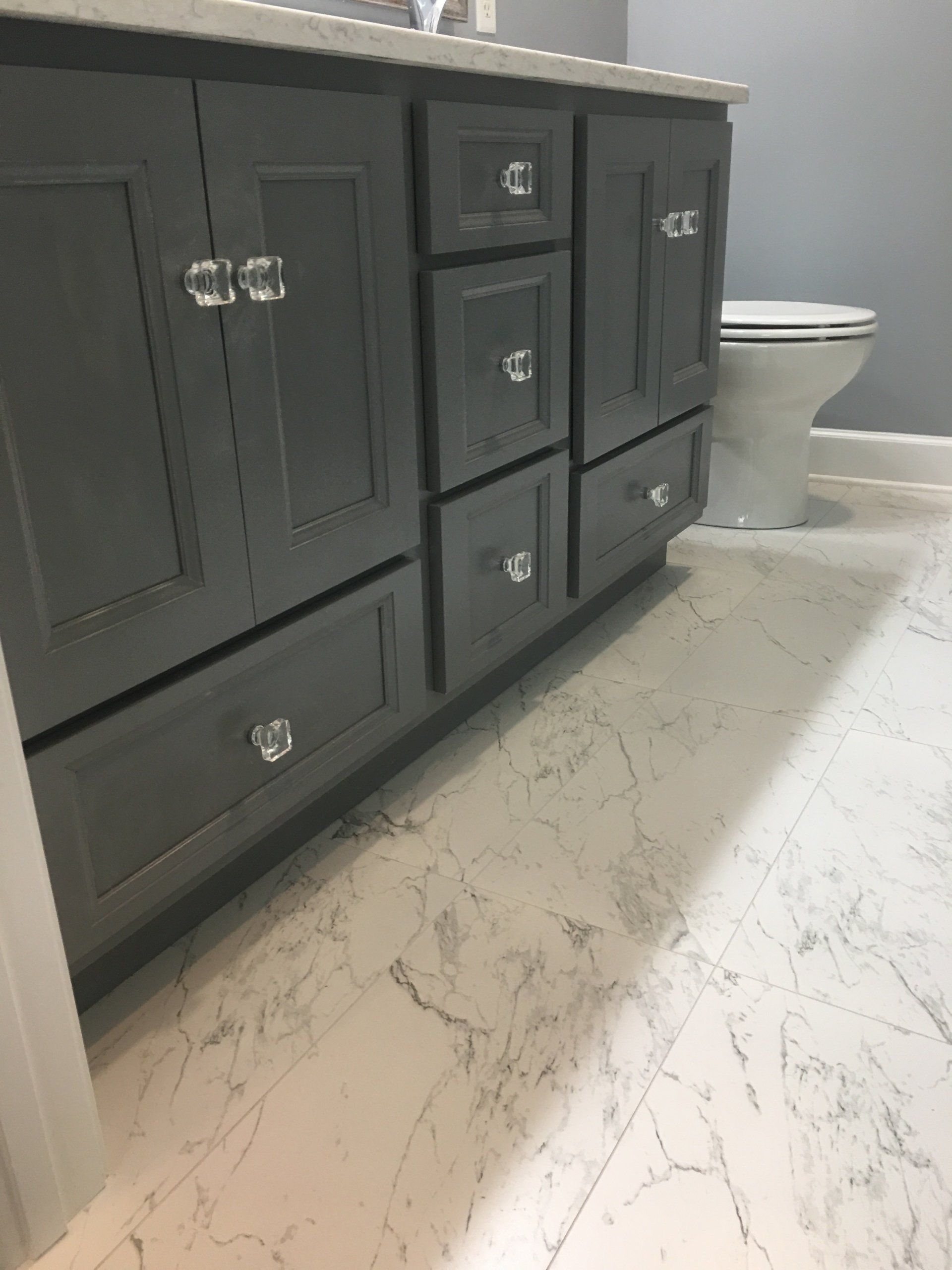Gray bathroom vanity with crystal knobs, white marble-look floor, and a toilet in the background.