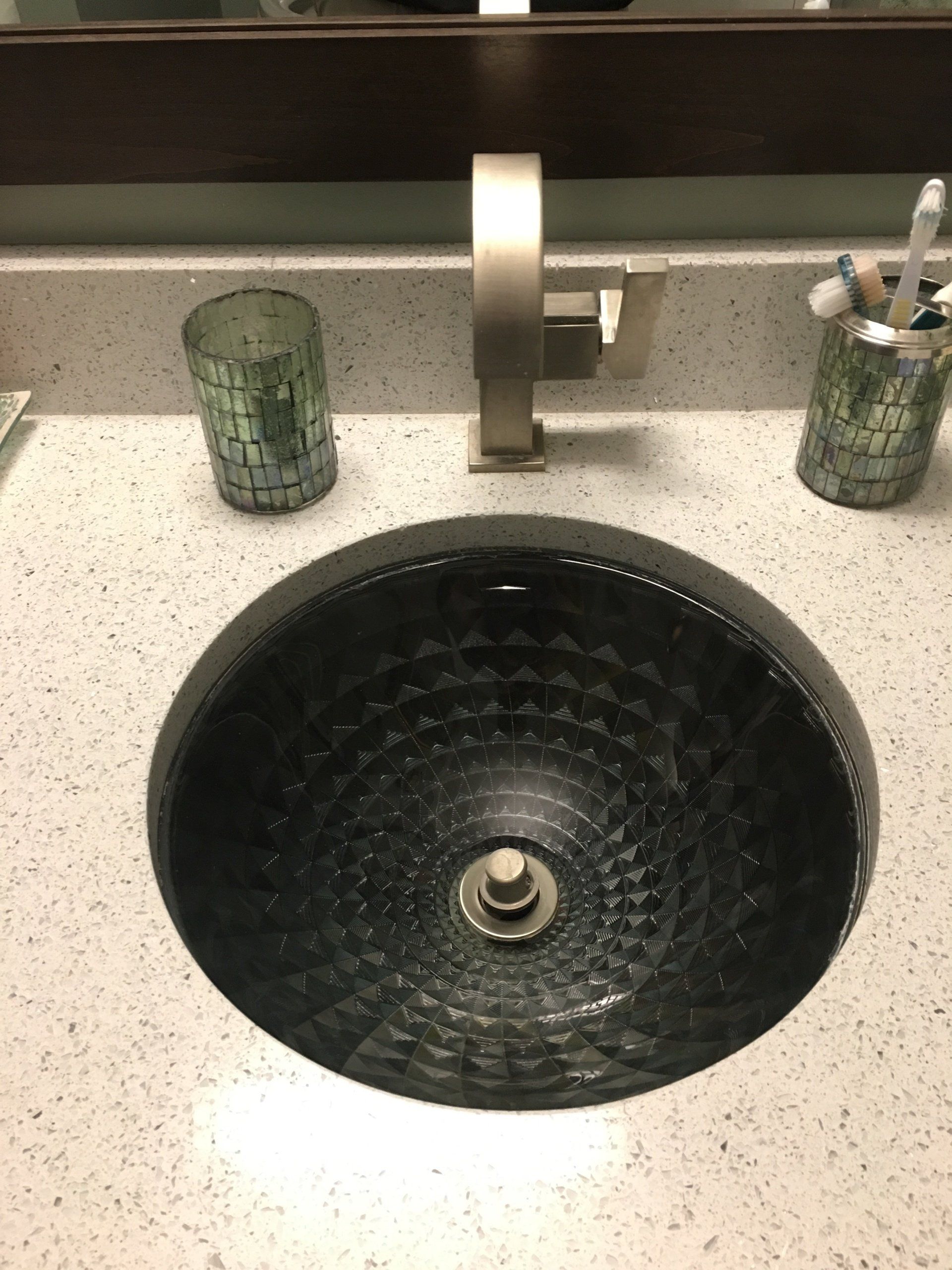 Black glass vessel sink on speckled countertop with silver faucet and green glass cup.