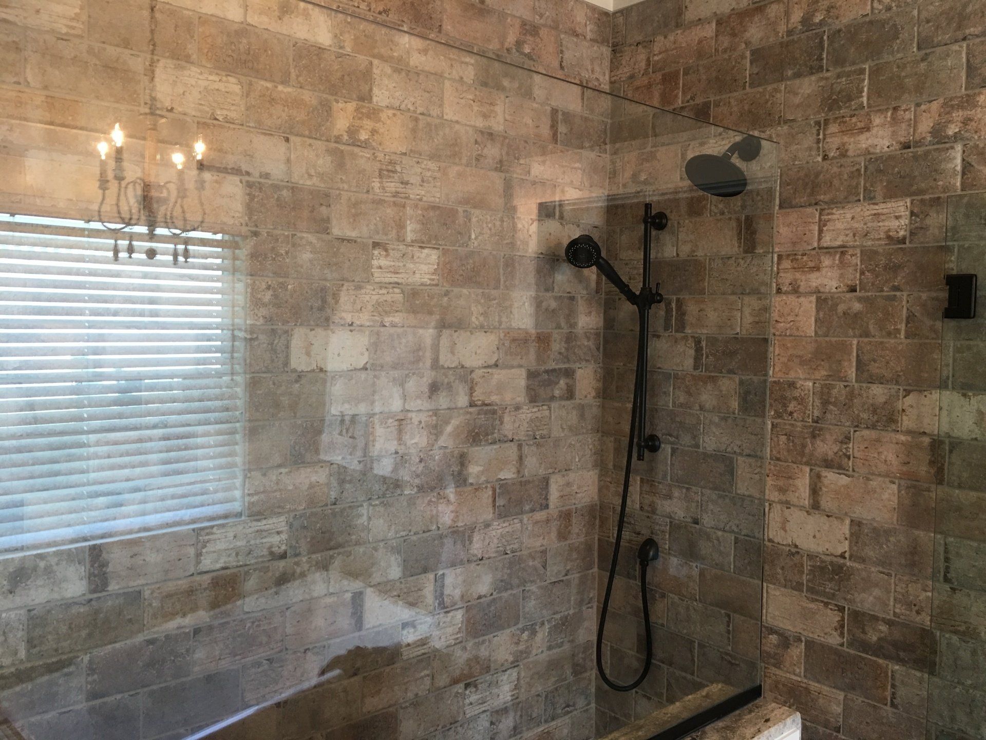 Shower with light brown brick-like tile walls. Black shower fixtures. Window with blinds.