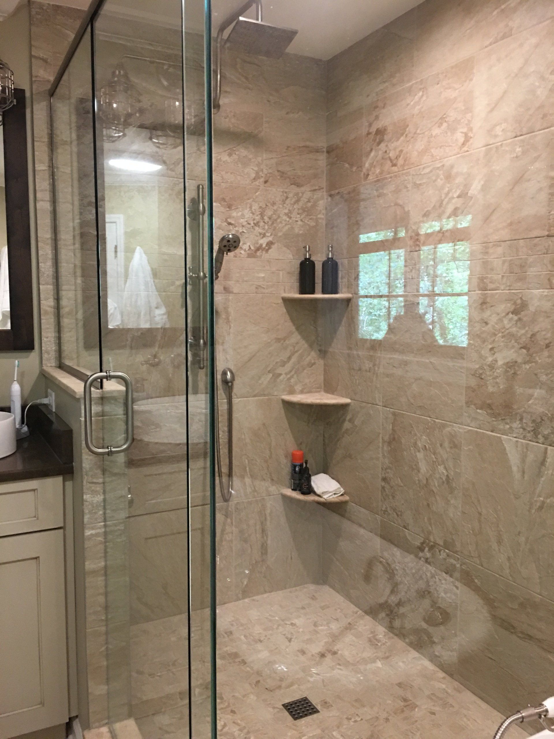 Bathroom with clear glass shower, tan tile walls, two shelves with toiletries.