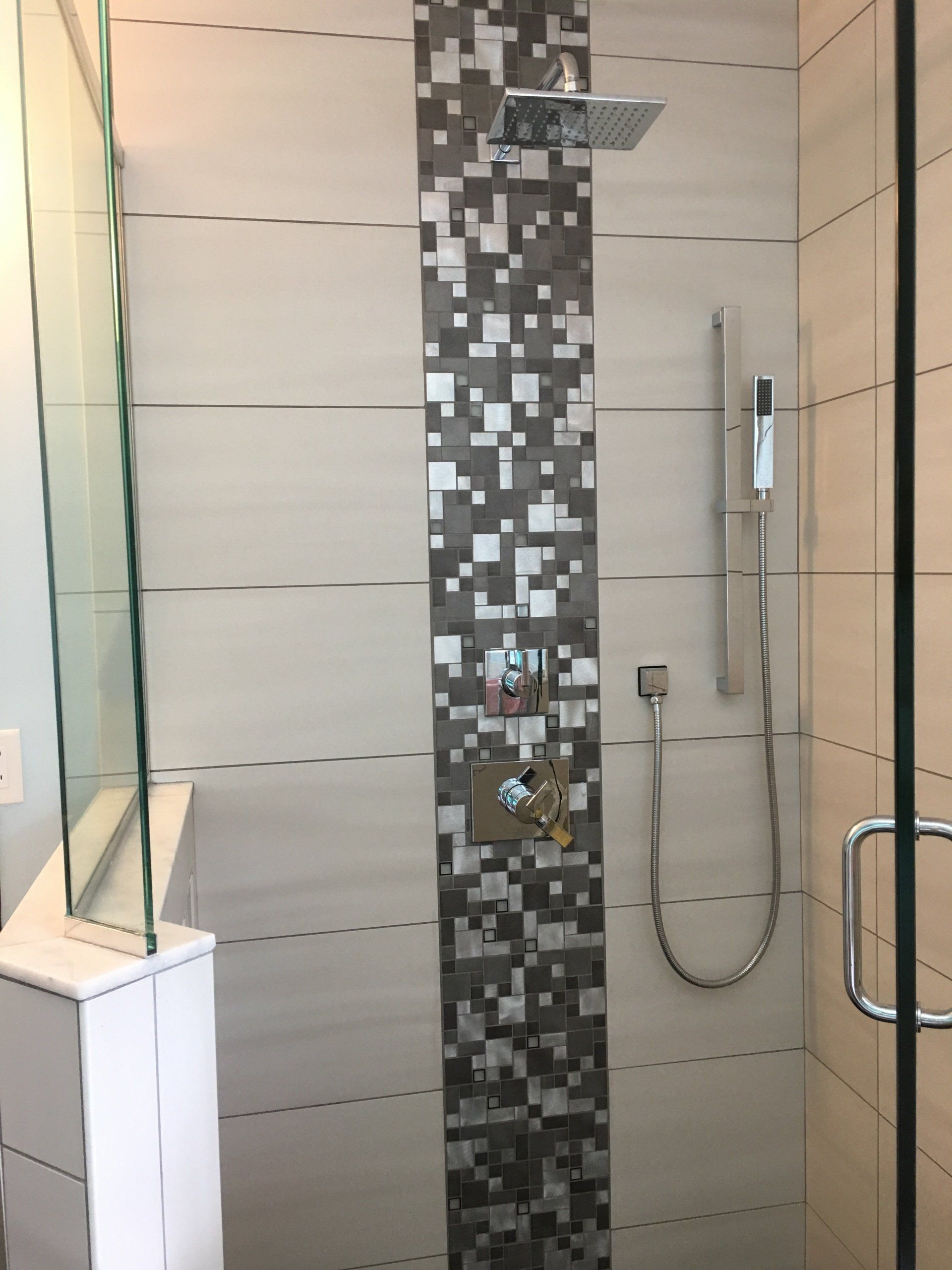 Shower stall with glass door, white and gray tiled walls, square showerhead and handheld sprayer.