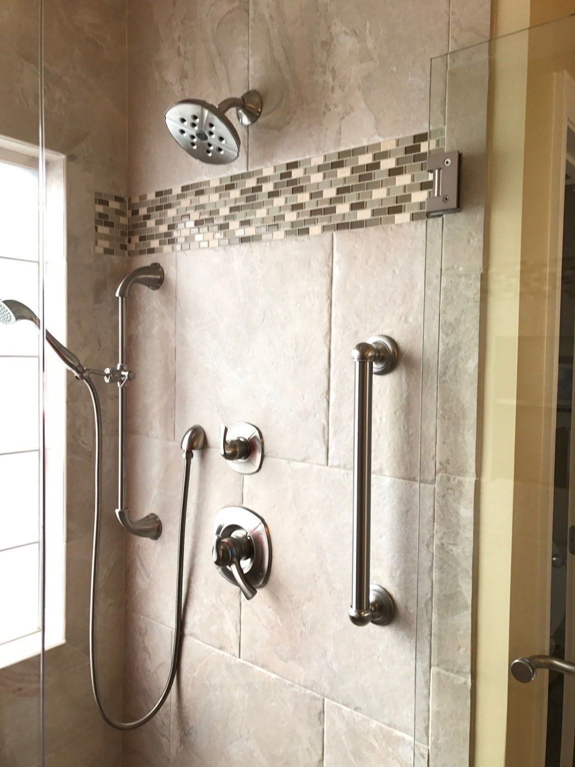 Shower with grab bar, multiple shower heads, mosaic tile accent, and glass door.