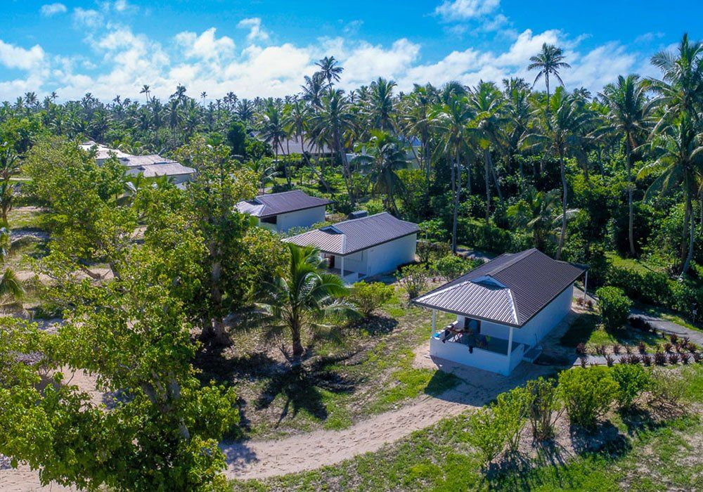 Small white houses with gray roofs along a dirt road in a lush tropical landscape under a blue sky.