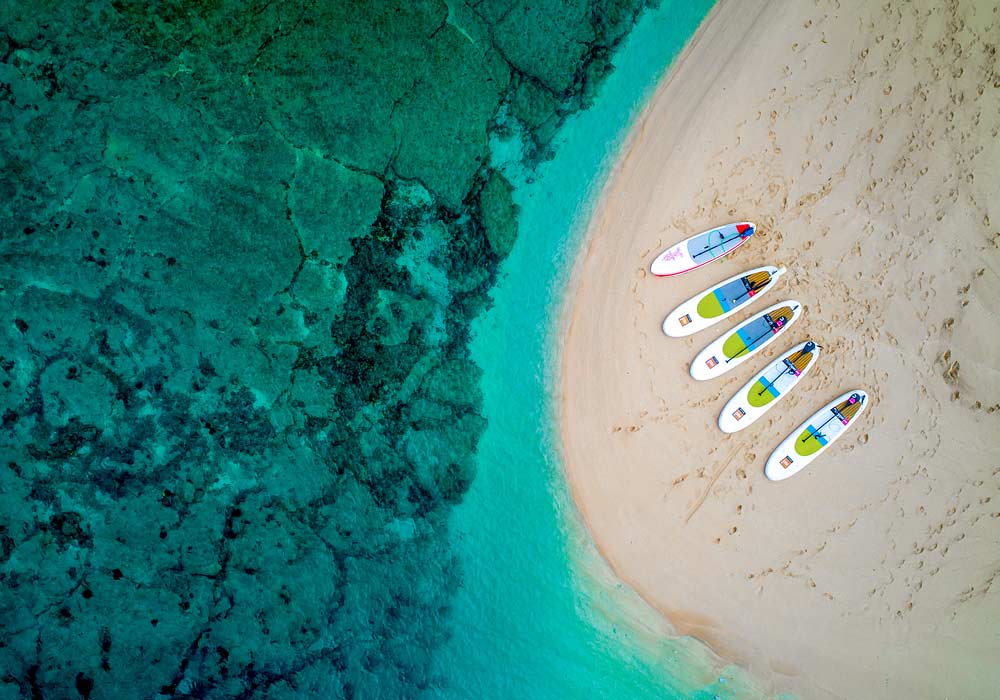 Aerial view of colorful surfboards on a sandy beach beside turquoise water.