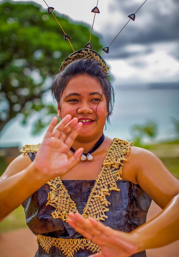 Person in a patterned dress smiling and waving outdoors near water with cloudy sky.