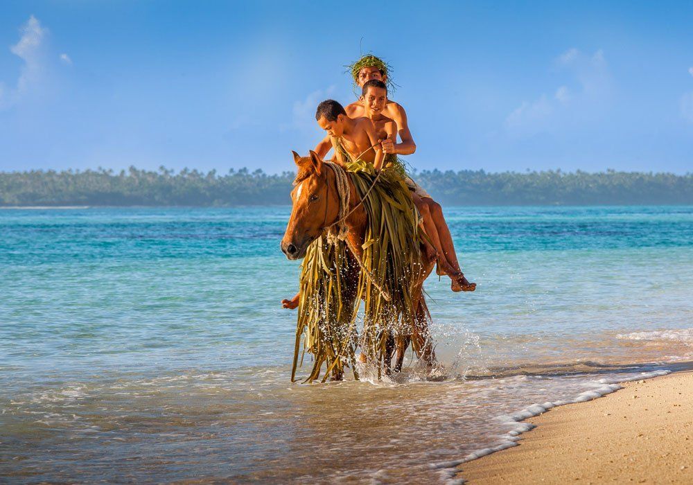 A person rides a horse through shallow ocean water near a sandy beach under a blue sky.