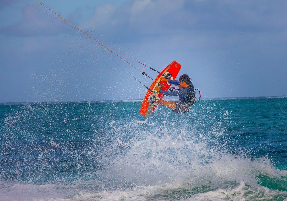 Kitesurfer airborne over ocean spray, holding a bright orange kiteboard under a blue sky.