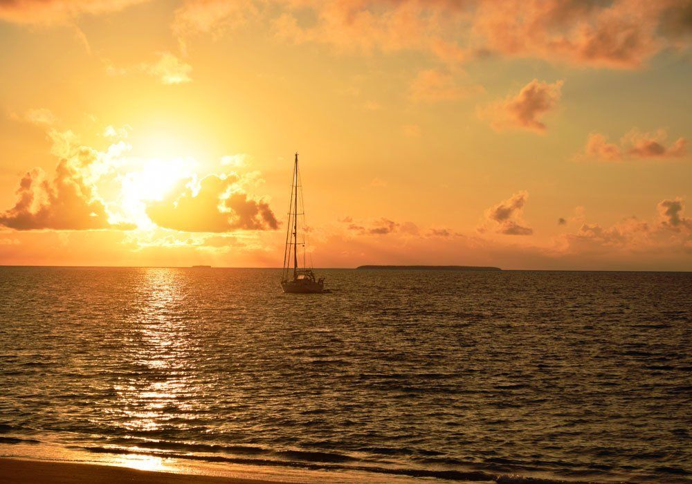 Sunset over calm ocean with a sailboat silhouetted near the horizon