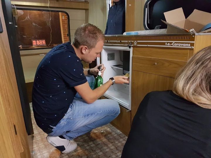 Een man opent een minikoelkast in een camper. Hij knielt bij de koelkast en houdt een groene fles vast.
