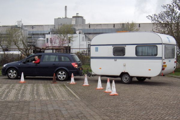 Een blauwe auto trekt een witte caravan door een parcours met kegels in een openluchtomgeving. De bestuurder zit in de auto, met een industriële achtergrond.