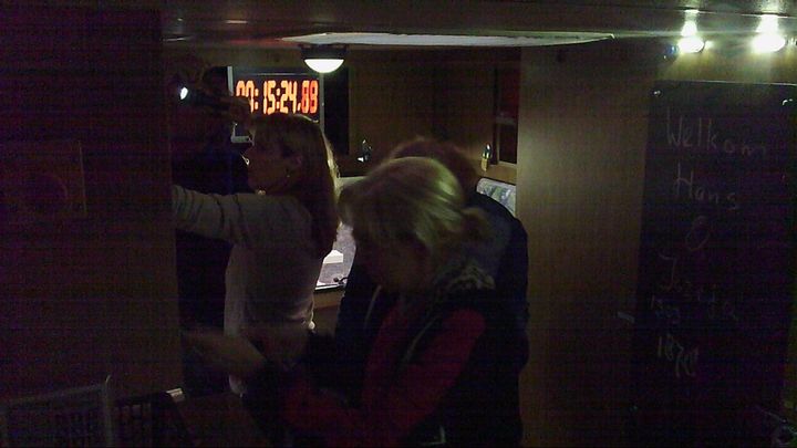 Mensen in een schemerig verlichte ruimte; een van hen met blond haar reikt naar iets aan de linkermuur, de ander staat met de rug naar de camera.