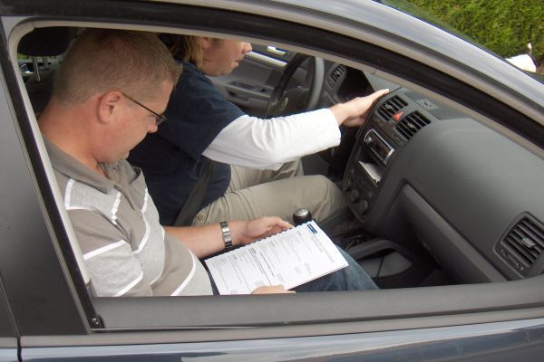 Een man zit achter het stuur en bekijkt documenten, een vrouw zit op de passagiersstoel en wijst naar het dashboard in de auto.