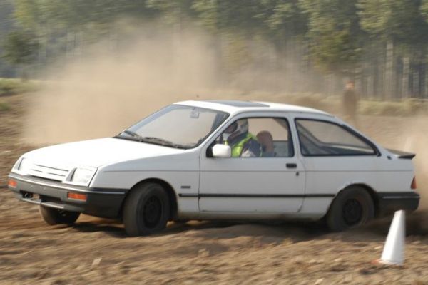 Een witte Ford Sierra drift over een onverharde weg en jaagt stof op.