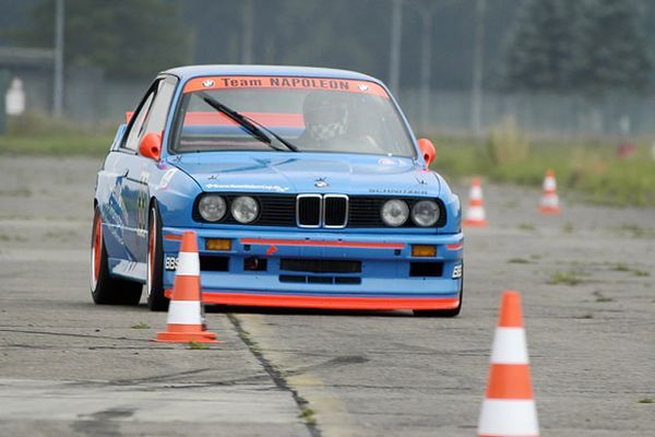 Een blauw-oranje BMW E30 raceauto rijdt over het circuit en nadert oranje kegels.
