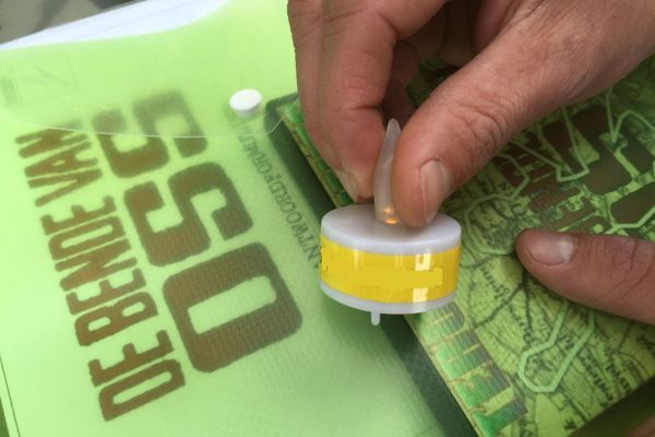 Een hand houdt een geel-wit plastic schijfvormig voorwerp vast boven een groen bedrukt oppervlak.