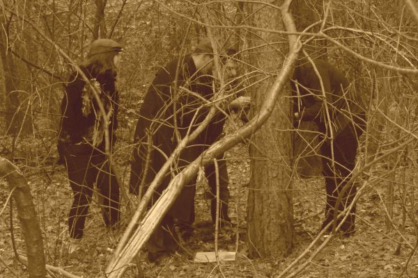 Drie figuren in donkere kleding, verborgen door takken, onderzoeken iets op de grond in een bosrijk gebied.