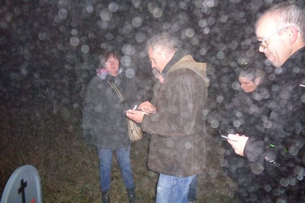 Mensen verzamelden zich 's nachts op een begraafplaats tijdens de regen. Eén persoon houdt een apparaat vast, mogelijk om een ​​meting te verrichten.