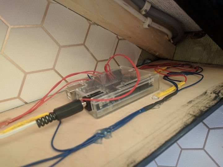 Close-up van een transparante doos met draden, verbonden met een zwarte kabel, op een houten ondergrond van witte zeshoekige tegels.